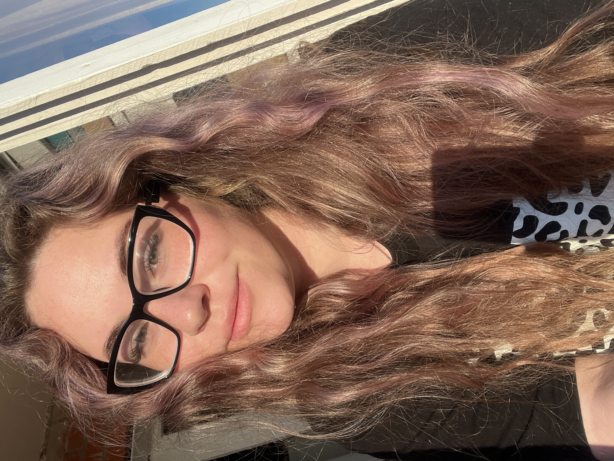 A young woman lies on her side, head near a window, with long, wavy, light brown hair with pinkish tones. She is wearing black-framed glasses. A window frame is visible in the upper left. She is wearing a dark-colored shirt with a white and black spotted pattern visible on the right side of the frame. The lighting suggests an indoor setting with natural light. There is no explicit indication of the location being Chaikovsky, Russia.