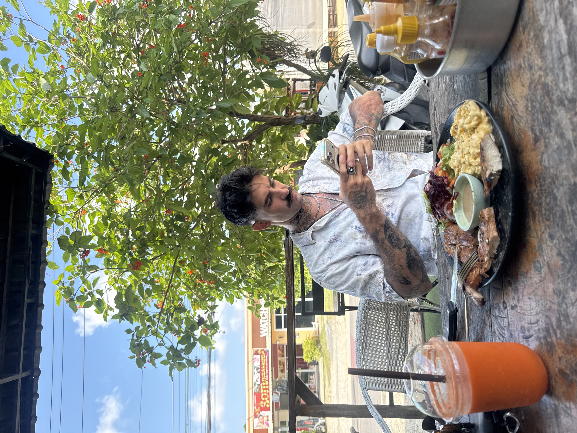 The image depicts a man with dark hair and tattoos, dressed in a white patterned shirt, sitting at a wooden table outdoors. He is holding a smartphone, seemingly taking a picture of his food. The table also holds a plate of food containing scrambled eggs, salad, and grilled meat, a glass of orange juice with a straw, and a container of sauces.
The scene is likely at a restaurant or cafe in Chalong, Thailand, based on the presence of buildings and businesses in the background, including one with the word "WATCH". A white scooter is also visible behind the man. A tree with green leaves and small red flowers provides shade. The sky is partly cloudy.