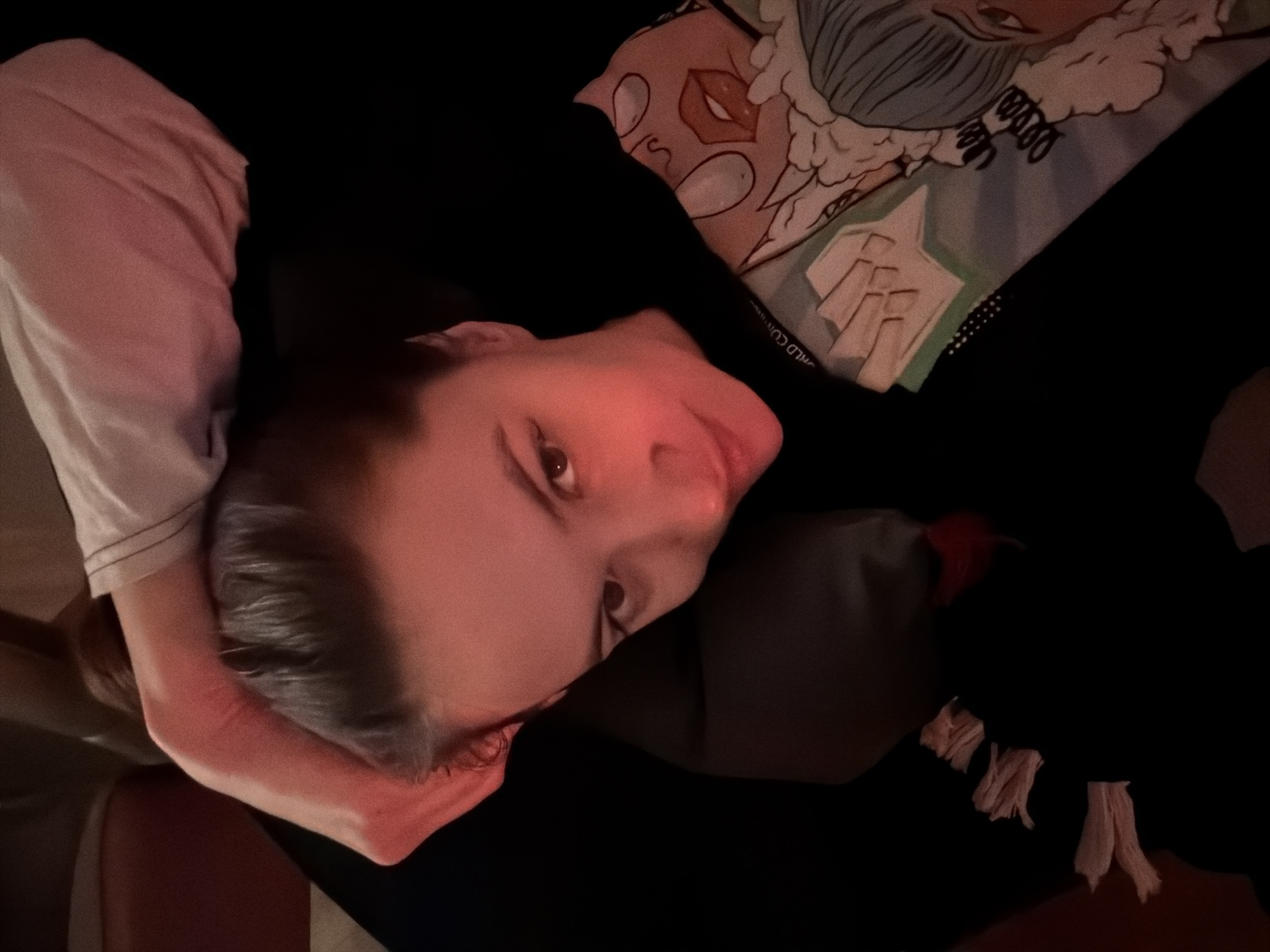 The image captures a person, likely an adult female, lying down and looking directly at the camera. Her head is supported by her left arm, which is clad in a white short-sleeved top. She is wearing a dark, possibly black, garment, which appears to be a hoodie or sweater, with faint white text "WORLD CO..." visible on it near her shoulder.
The setting is indoors and dimly lit, suggesting evening or night, with a warm, reddish artificial light illuminating her face and arm. She rests on a dark surface, possibly a couch or bed, with a decorative pillow positioned behind her head. The pillow features an anime or cartoon-style illustration, including a character with light blue hair and a separate graphic of red lips. The person's expression is neutral and calm, with a direct gaze towards the viewer. Tassel-like elements are visible on the lower right side of the image, potentially part of a blanket or the person's clothing. The overall atmosphere is relaxed and intimate. Based on the provided context, the location is Belgrade, Serbia.