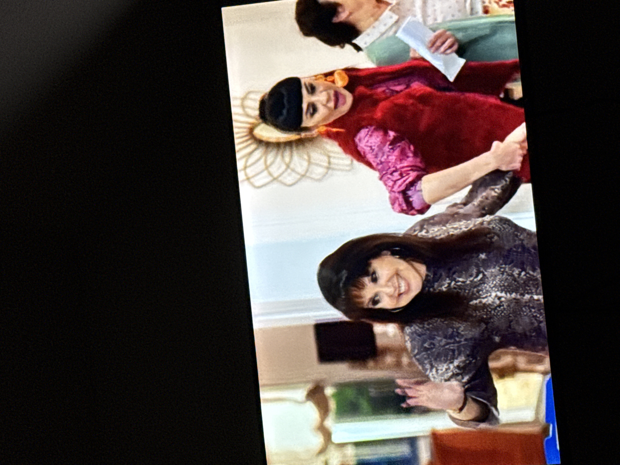 The image captures a scene with three women indoors. The woman on the left is smiling and waving with her left hand, wearing a long-sleeved, patterned top. The central woman has dark hair with bangs, large orange earrings, and is wearing a bright pink, puffy-sleeved top with a red vest. She is holding a piece of paper in her right hand and her left hand is clasped with the woman on the right, who is wearing a light green shirt. The background shows interior details like curtains, a dark rectangular object on the wall, and a decorative wall hanging with a circular pattern. The scene is brightly lit. The location is not specified within the image.