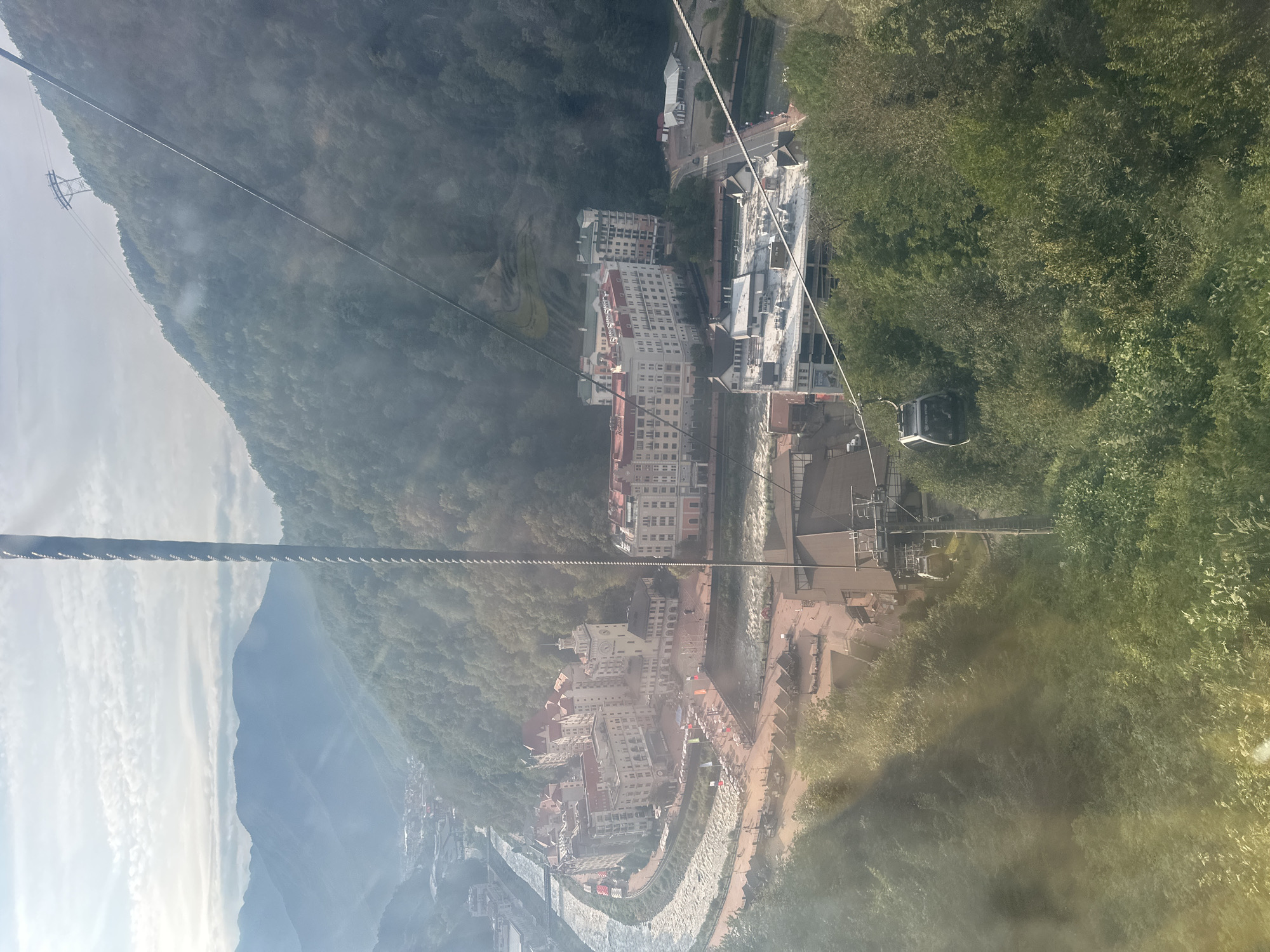 This is an aerial shot from a cable car in Estosadok, Russia. The view looks down on a resort town nestled in a mountainous valley. A winding river is visible, with several large, multi-story buildings lining its banks. These buildings appear to be hotels or resorts, characterized by their light-colored facades and red or beige roofs. In the foreground, a cable car ascends a steep, tree-covered mountain. The sky is partly cloudy, with sunlight filtering through the clouds and casting shadows across the landscape. The overall impression is one of a picturesque mountain resort town.
