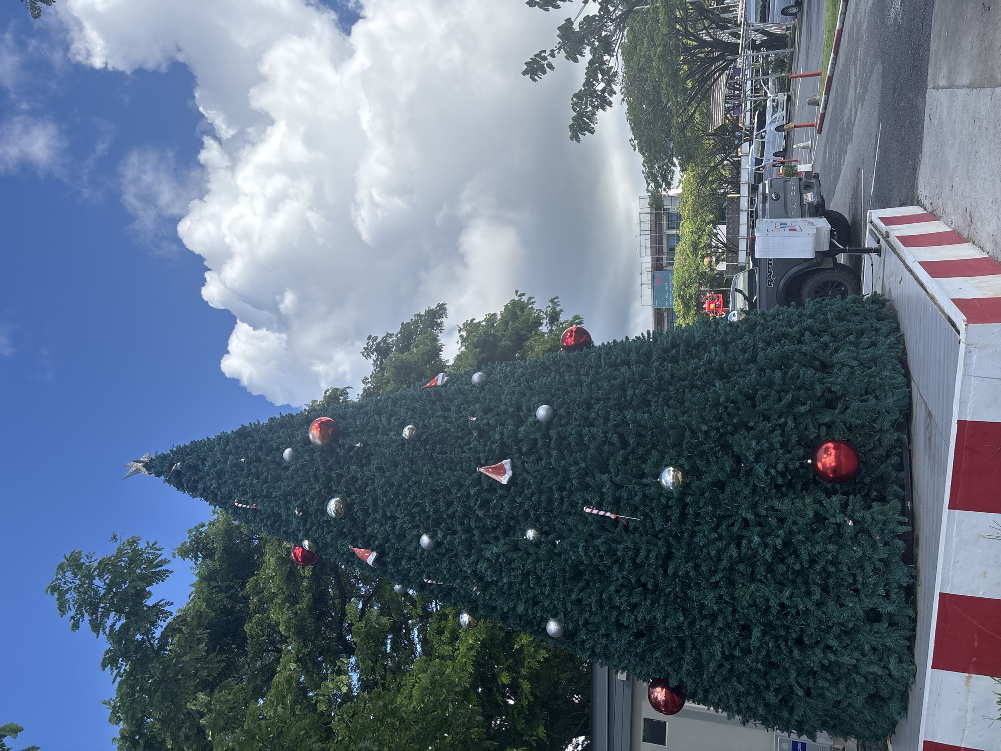 The image depicts a large, artificial Christmas tree decorated with red and silver ornaments, including some resembling candy canes and Santa hats. A star sits at the very top of the tree. The tree is situated outdoors, with lush green foliage from other trees framing it from below and to the left. In the background, a paved road is visible, lined with cars and a utility trailer attached to a dark-colored truck. A building with a billboard is also in the distance. The sky above is a mix of bright blue and dramatic white clouds, suggesting a partly cloudy day. The lighting indicates it is daytime. The scene appears to be set in a public area, possibly near a road or a park. There is no indication of people or specific events taking place, beyond the display of the decorated Christmas tree. Visible text includes "RAPTOR" on the truck and "Haier" on a label on a white box on the trailer. The red and white striped barriers along the right side of the image suggest a temporary road closure or a construction zone.