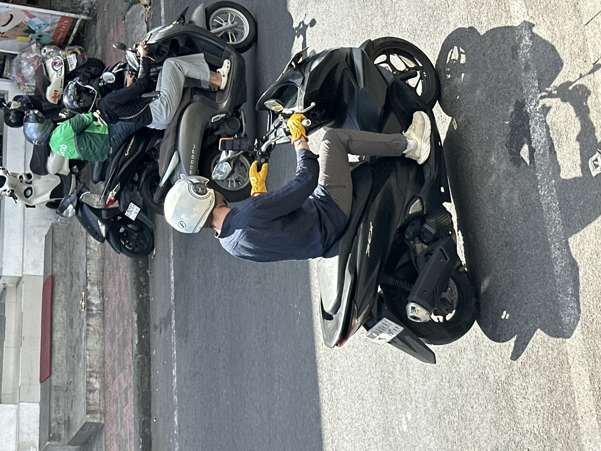 The image shows a street scene in Ubud District, Indonesia, with several motorbikes. In the foreground, a person wearing a dark blue long-sleeved shirt, grey pants, white socks, and light-colored shoes is riding a black scooter. They are wearing a white helmet and yellow gloves. A mobile phone is mounted on the scooter's handlebars. Behind this rider, another scooter is visible with a person wearing a black hoodie and grey pants, also wearing a helmet. Further back, a motorbike with a rider in a green jacket with "Grab" visible on it is parked, along with other parked motorbikes. The street is paved, and a sidewalk with red bricks and concrete steps is visible to the left. The lighting suggests it is daytime, with shadows cast by the motorbikes and riders on the asphalt.