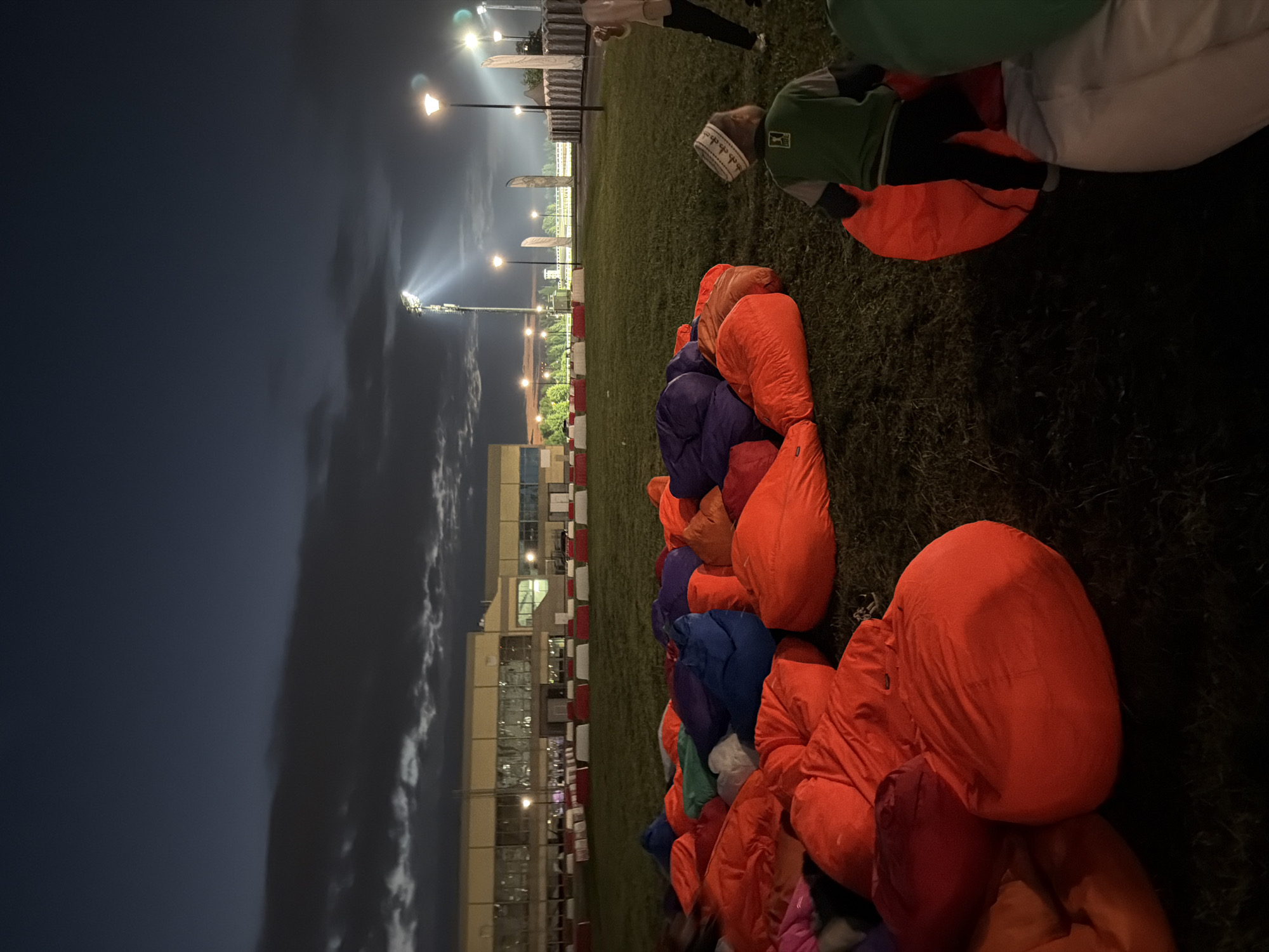 A group of beanbag chairs are scattered on a grassy field at dusk. The chairs are various colors including orange, blue, purple, and green. In the background, a modern building with large windows is visible, along with several tall light poles that are illuminated, casting beams of light into the night sky. A small child wearing a green shirt and a white kufi is seen in the foreground, near a large orange beanbag chair. Another person, partially visible on the right side of the frame, is standing near the child. The overall scene suggests an outdoor event or gathering.