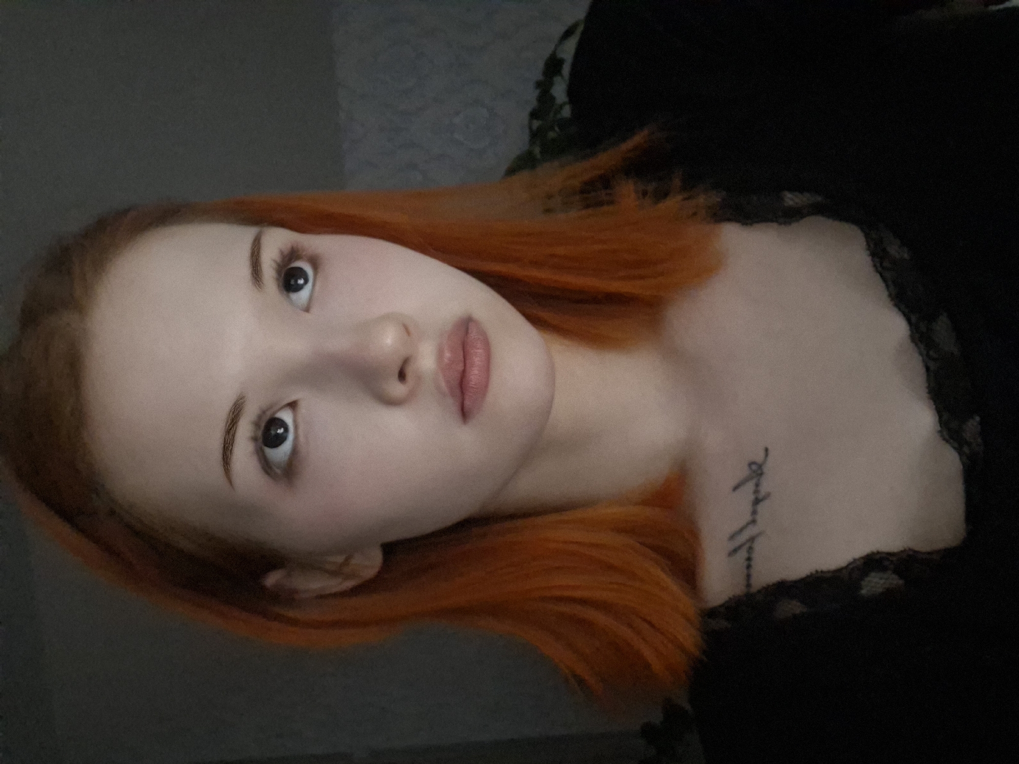 The image depicts a fair-skinned individual with orange hair. The person's face is angled towards the camera. They have light eyebrows, dark eyes, a small nose, and light pink lips. The individual is wearing a black garment with a lace trim. A tattoo is visible on the upper chest. The background appears to be a dark gray wall, with a plant partially visible in the upper right corner. There is no indication of this being located in the city of Chaikovsky, Russia, or any other specific location.