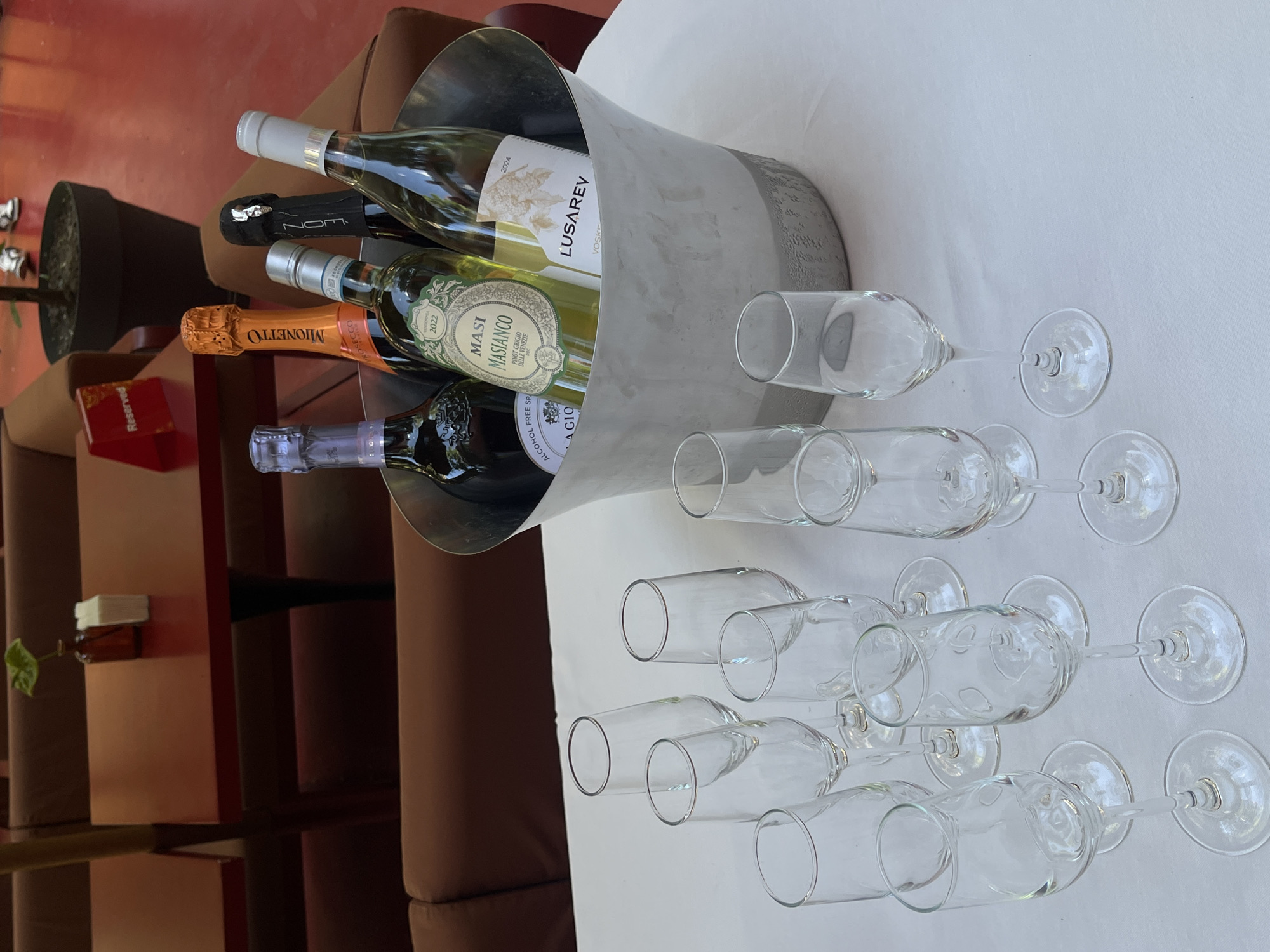 The image shows a collection of wine bottles in an ice bucket and arranged champagne glasses on a white tablecloth, likely in a restaurant. Four wine bottles are visible in the bucket: LUSAREV VOSKE (2024), MASI MASIANCO PINOT GRIGIO DELLE VENEZIE (2022), MIONET PROSECCO, and a bottle labeled "NOM ALCOHOL FREE SP A GIO." There are ten champagne glasses arranged in two rows on the tablecloth. "Reserved" card visible on a table with a brown surface and red trim. The setting is indoors with brown furniture and possibly located in Yerevan, Armenia.