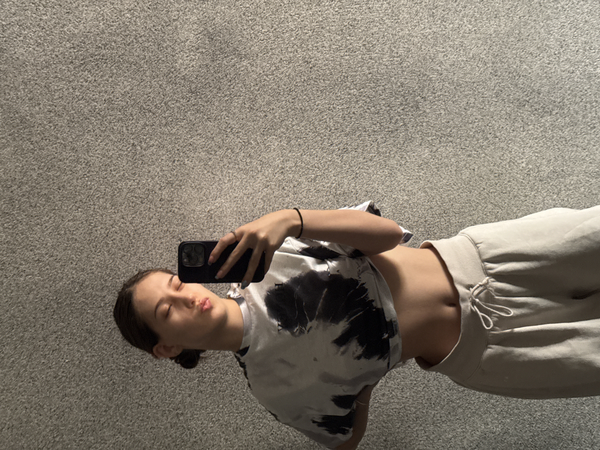 A casual selfie captures a young woman posing indoors in what appears to be a residence in Sofia, Bulgaria. The image is rotated 90 degrees counter-clockwise, showing the woman positioned horizontally against a grey, speckled carpet floor. She is holding a dark-colored smartphone, possibly an iPhone, in her left hand, using it to take the photo. Her head is tilted slightly, and her eyes are closed, with her lips pursed in a playful "kiss face" expression.
The woman has dark hair pulled back. She is wearing a white crop top with a distinct black tie-dye pattern, revealing her midriff. Her lower body is clad in light beige or grey sweatpants/joggers with a visible drawstring at the waist. Accessories include a simple black string bracelet on her right wrist and a ring on the ring finger of her left hand. The background consists solely of the consistent texture of the light grey carpet, suggesting a clean, uncluttered indoor environment. The lighting appears soft and even, indicating an indoor shot without harsh shadows. No visible text is discernible on her clothing or the phone.