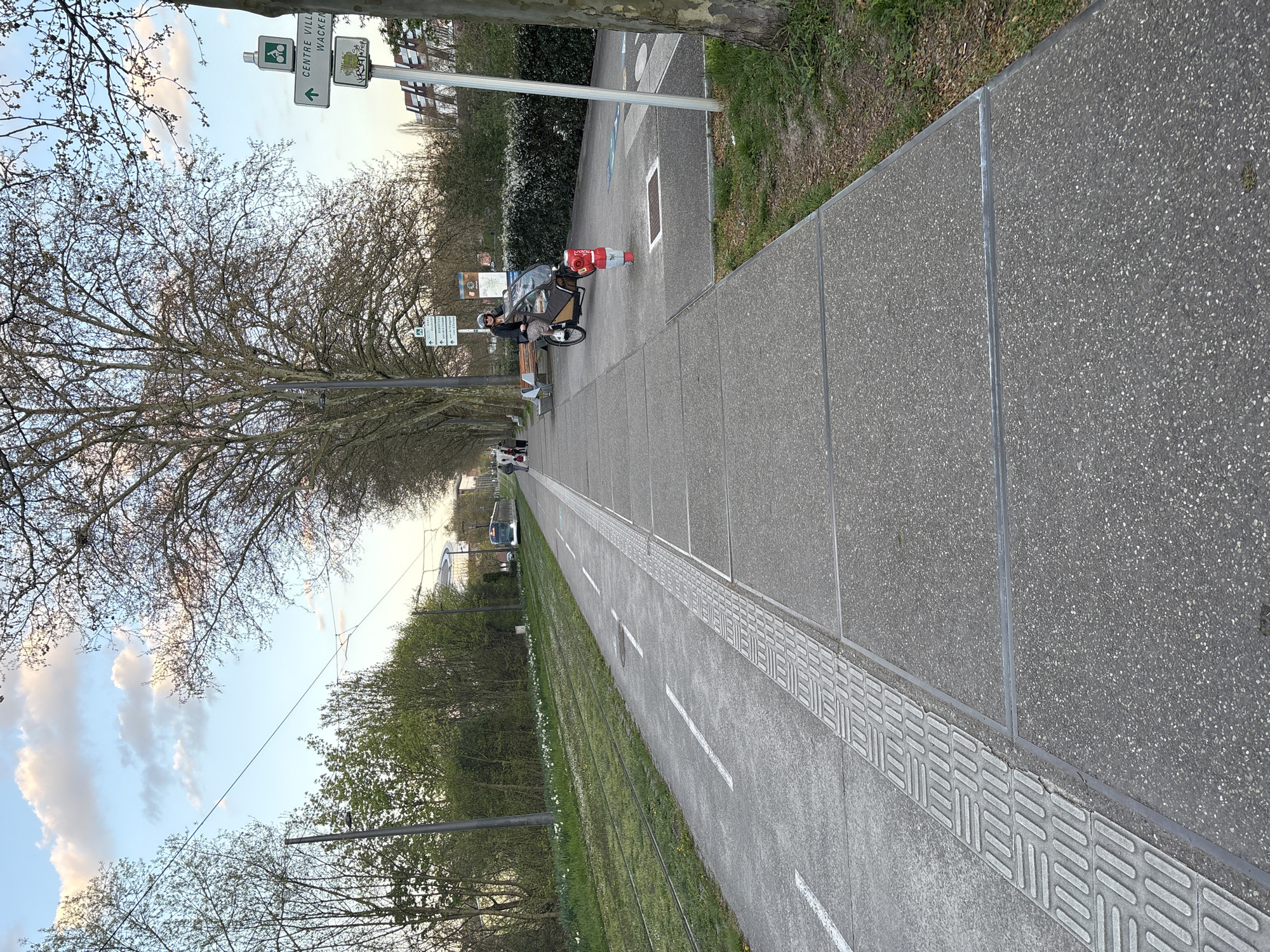 The image shows a long, straight paved path, likely a section of a tram line, stretching into the distance. On the left side of the path is a strip of grass with white flowers blooming. Overhead tram wires run parallel to the path. A person wearing a helmet is seen sitting on a bench next to a three-wheeled cargo bike. The cargo bike contains what appears to be a wrapped-up item, possibly a large package. Signage indicating directions is visible, one of the signs saying "Centre Ville Wacken." The location appears to be in Strasbourg, France, specifically near the Wacken area, a business and exhibition district.
