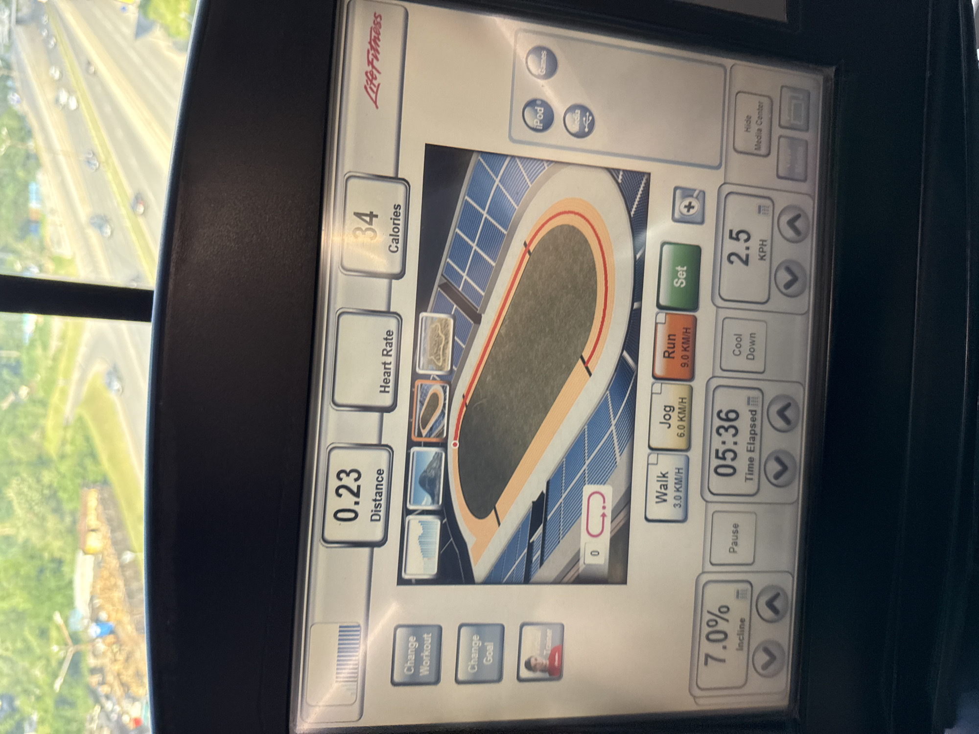 The image shows the display screen of a Life Fitness treadmill. The display indicates that the user has walked a distance of 0.23 (units not specified, presumed miles or kilometers), burned 34 calories, and has been exercising for 5 minutes and 36 seconds. The treadmill is set to a 7% incline and a speed of 2.5 KPH. The treadmill also displays a graphical representation of a running track. Through a window behind the treadmill, a blurred view of a city can be seen. Though I cannot discern the city of Sofia, Bulgaria, based on the visible image information.