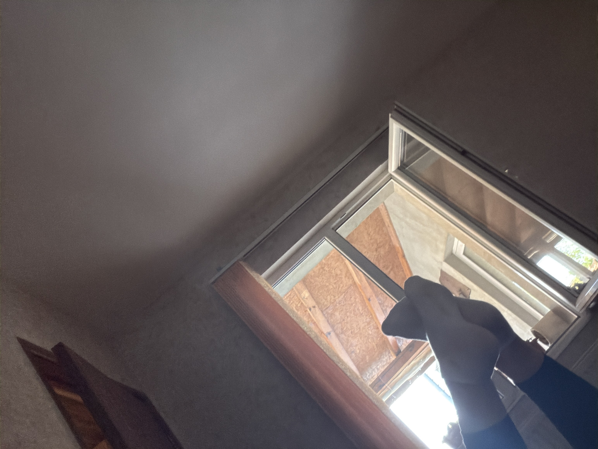 The image captures an interior view, dominated by a brightly lit window area. In the lower right, a person's hands are visible, wearing light-colored gloves and dark sleeves. The hands appear to be interacting with the window, possibly opening it further, adjusting something, or cleaning.
The window itself is partially open, revealing a clear, bright daytime outside. What is visible beyond the window suggests a construction or renovation site, with exposed plywood or OSB sheeting and wooden framing. The window frame is a light color, likely white or silver. Below the window is a sill or frame constructed from a light-colored, possibly particle board, material with a reddish-brown finished edge.
The room's ceiling, stretching across the upper left and center, is a light grey color. The wall in the lower left corner has a subtle texture, perhaps wallpaper, and a dark, possibly wooden, object or door frame is partially visible there. The interior of the room appears somewhat dim in contrast to the strong light flooding in from the window. No text is visible in the image.