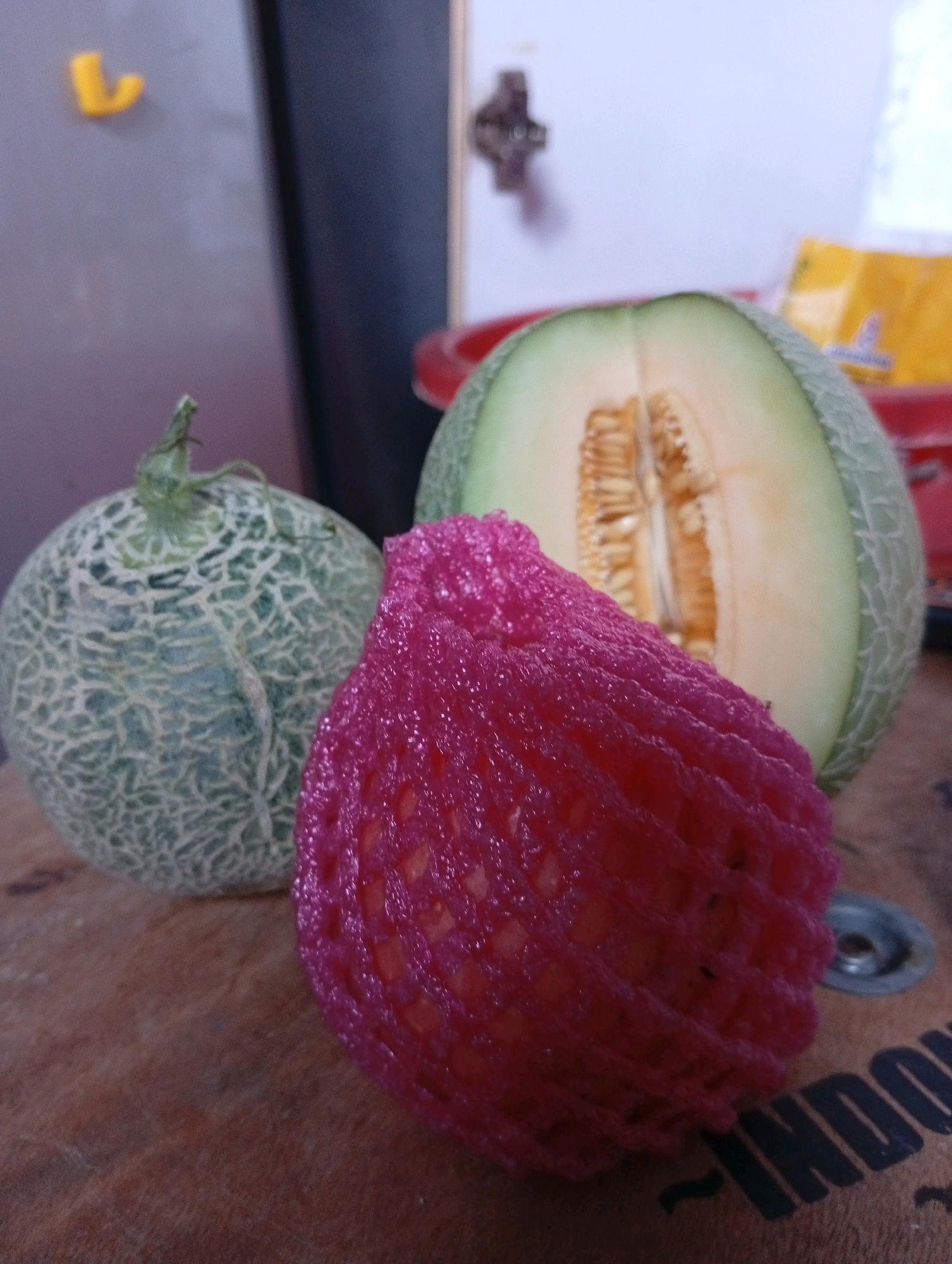 The image shows a close-up of three fruits on a wooden surface. On the left is a whole cantaloupe with a net-like pattern on its rind. In the center, also in its entirety, is another cantaloupe, this one wrapped in a pink mesh net. To the right, a cantaloupe has been cut in half, revealing its orange flesh and seeds. The background is blurred, but hints of a kitchen environment are visible, including a refrigerator or cabinet and possibly some packaging. The lighting appears to be artificial, creating soft shadows. The scene suggests a domestic setting, possibly in the city of Kebondalem, Indonesia, where fresh produce is prepared or displayed. There are no people or actions visible in the image.