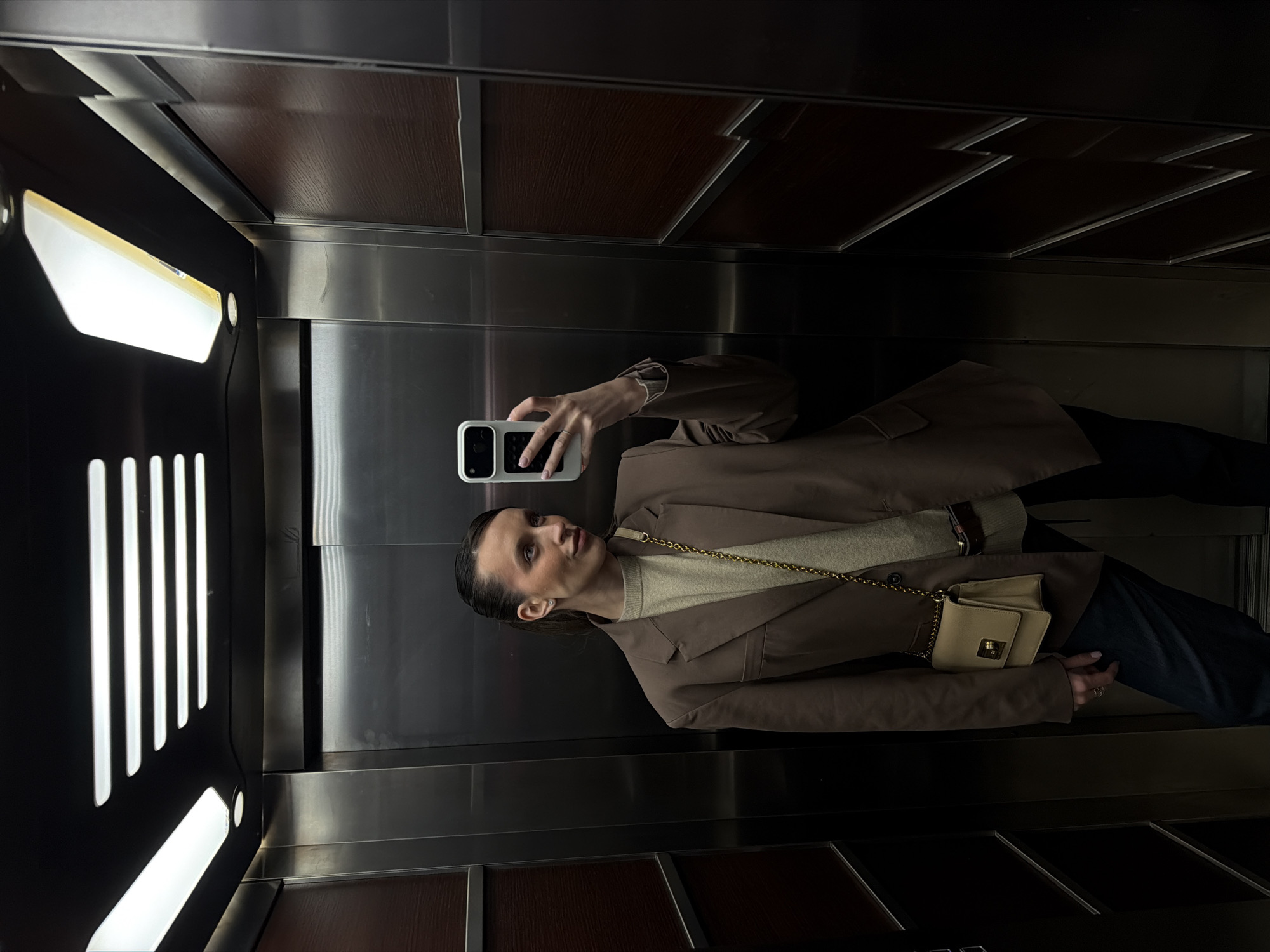 A woman is taking a selfie in an elevator. The elevator has brushed metal walls with dark wood paneling at the top and bottom. The lighting panel on the left side of the elevator has vertical illuminated slits. The woman is wearing a brown blazer, a light sweater, and dark pants, and she has a small, light-colored purse with a gold chain. She is holding her phone up to take a picture of herself in the elevator mirror. The city of Budva, Montenegro is not visible in the image.