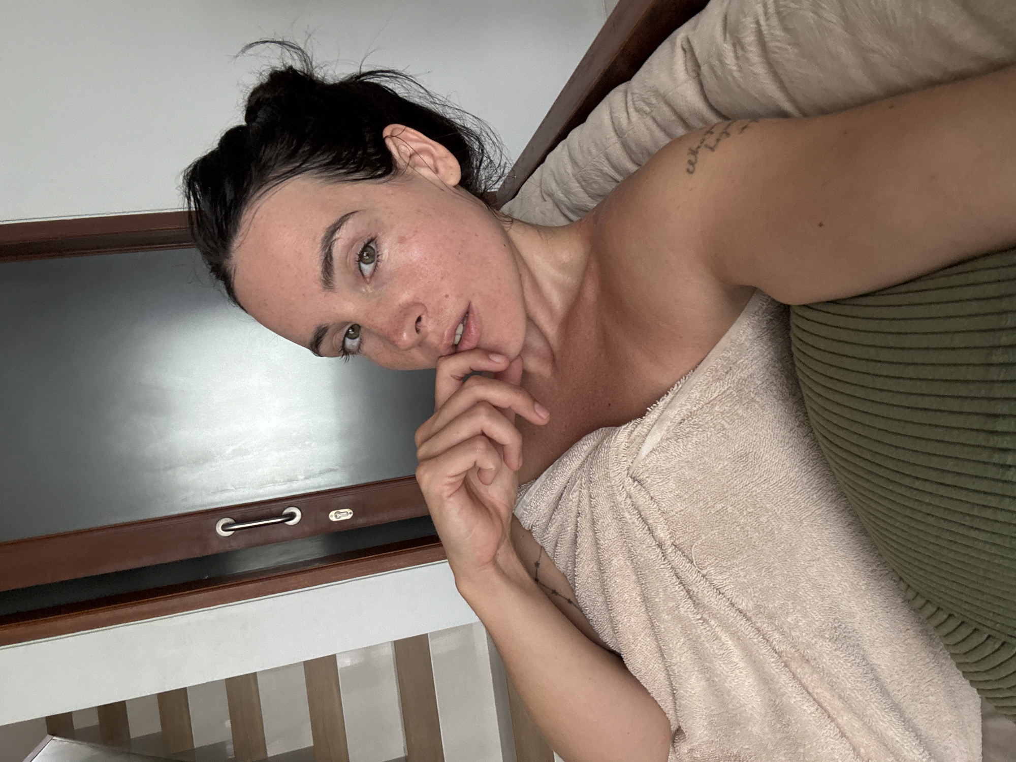 A young Caucasian woman with her hair pulled up in a bun is lying down, wrapped in a light brown towel. She is indoors, possibly in a bedroom or living area, with a wooden door and a staircase visible in the background. The woman is looking at the camera with a relaxed expression, her left hand resting on her chin. A tattoo is visible on her left shoulder. The lighting is soft, suggesting it could be natural light from a window.