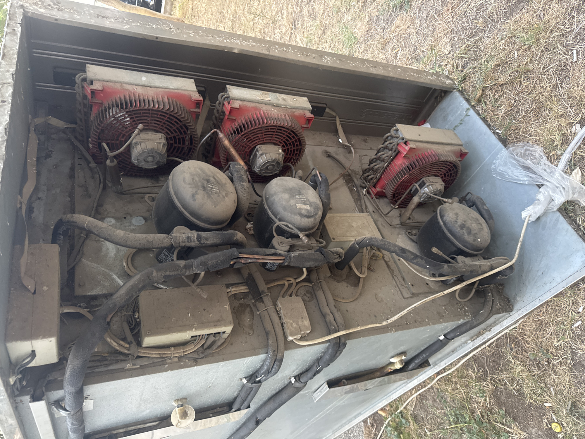 This is a close-up, forensic, matter-of-fact image of the internal components of an outdoor HVAC unit, likely a condenser, in Sutomore, Montenegro. The unit is partially open, revealing a complex arrangement of compressors, fans, and copper tubing, all heavily coated in dust and grime. Three large, cylindrical compressors are stacked vertically. Above and between them are three red-framed condenser fan assemblies, each with a large, circular fan. Numerous black insulated tubes and wires snake throughout the unit. The exterior casing appears to be weathered metal. The unit is situated outdoors on a patch of dry, sparse grass and dirt. A crumpled white plastic bag is visible on the ground near the base of the unit. The overall impression is one of disuse and neglect of the machinery.
