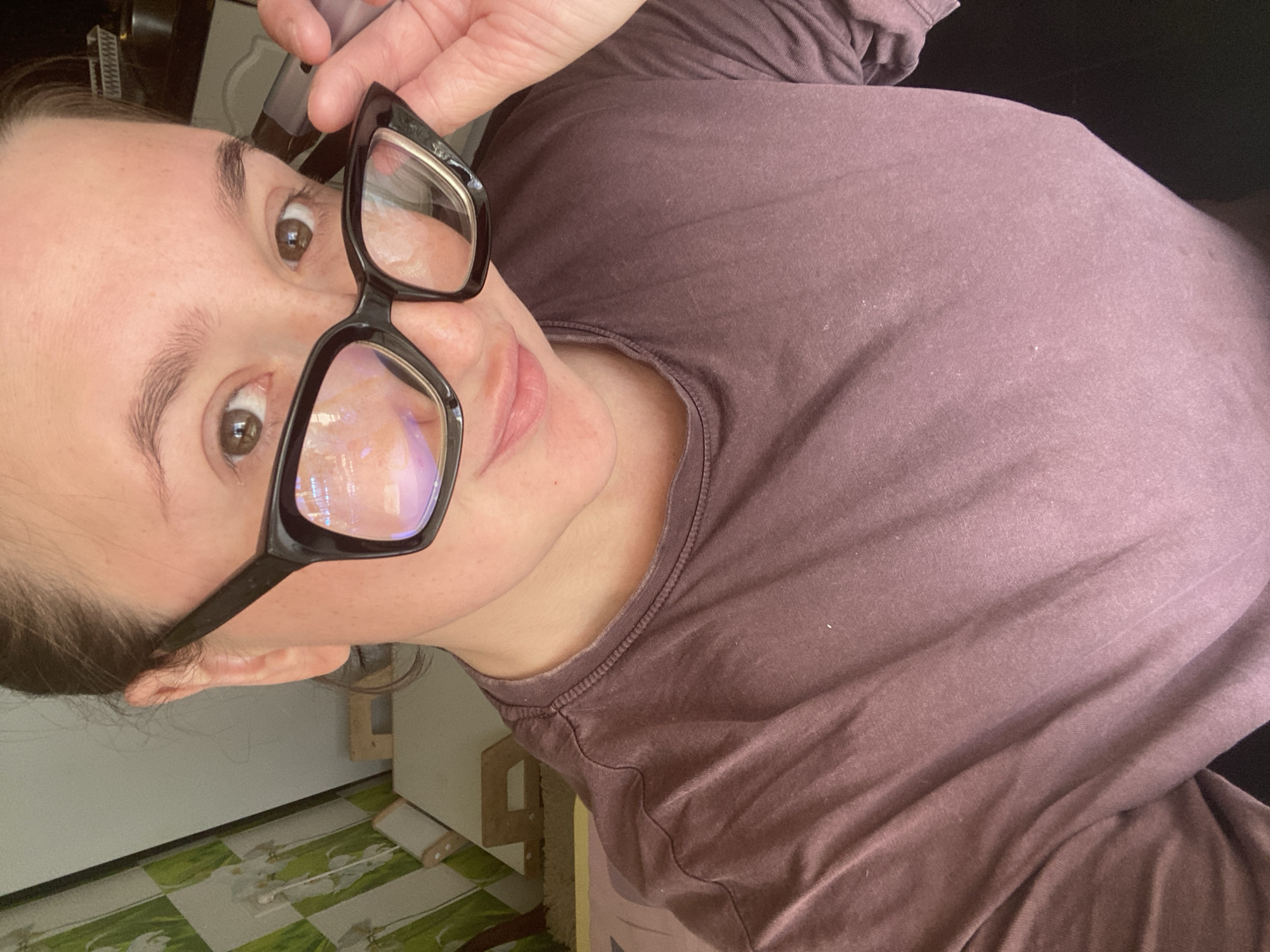 A woman, wearing a dusty rose colored t-shirt and large black framed glasses, is taking a selfie. She is holding the right side of her glasses with her right hand, and a silver electronic cigarette with her left hand. The background appears to be a domestic setting. To the left of the woman, a wall is visible with a light green wallpaper featuring a floral pattern, possibly depicting orchids. Next to the wallpaper, a wooden easel and a white canvas are visible. Further into the background, a shelf with white containers and a stainless steel appliance, possibly a kettle, can be seen. The lighting suggests an indoor environment, possibly with natural light coming from the left.