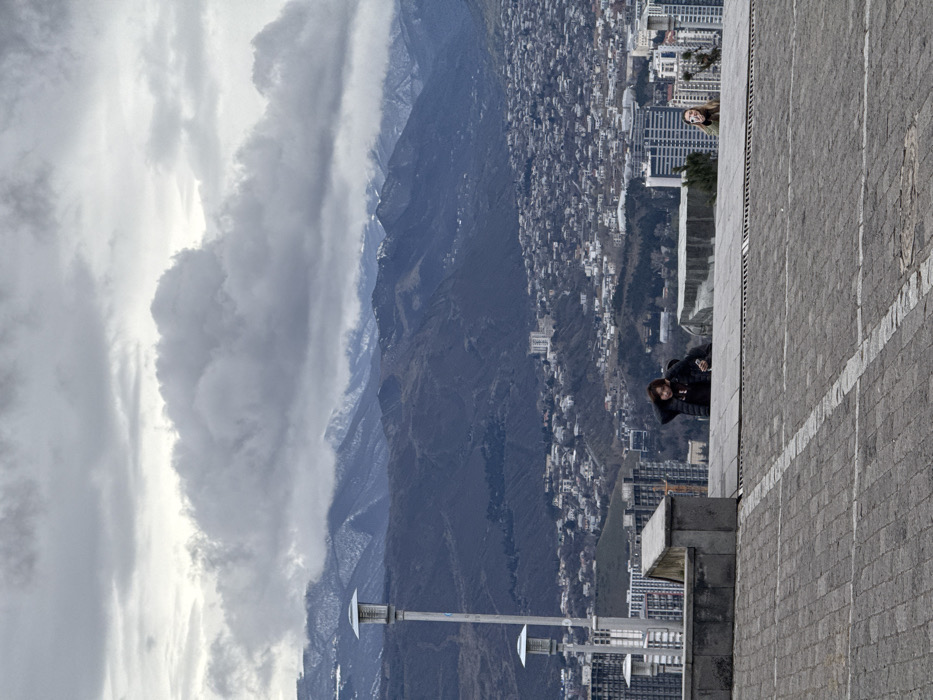 The image captures a panoramic view from a high vantage point overlooking the city of Tbilisi, Georgia, with distant mountains under an overcast sky. The sky is dominated by heavy, grey cumulus clouds. In the far background, dark, rugged mountains are visible, with patches of snow on their upper slopes and peaks. The mid-ground shows a vast urban sprawl of Tbilisi, featuring numerous buildings of varying heights and densities, including a visible cable car line.
In the immediate foreground, a grey paved surface, constructed of pavers with faint white grid lines, extends towards a low, grey concrete retaining wall. To the left of the frame, behind this wall, three tall, slender streetlights with multi-tiered circular fixtures are present. Two individuals are positioned behind the concrete wall, viewing the scene. On the left, a person with dark hair, wearing a dark jacket, has their left hand raised near their head and is holding a white object in their right hand, appearing to observe the distant cityscape. To the right, another individual with lighter hair, wearing a dark top with a light-colored design, smiles directly towards the camera.