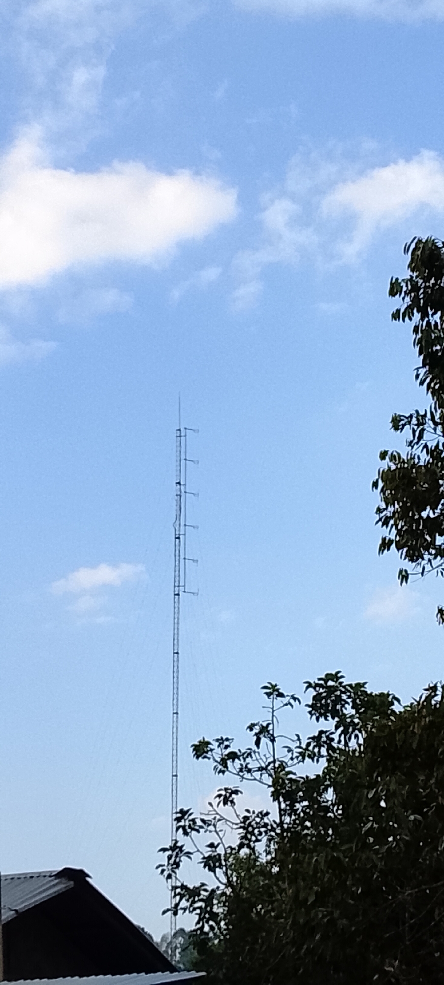 In the city of Padang Dalom, Indonesia, a tall radio tower stands against a bright blue sky with scattered white clouds. The tower is a slender, metallic structure with horizontal supports at intervals, extending upwards to a pointed antenna. To the right of the tower, a dense canopy of dark green leaves from a large tree partially obscures the sky. In the lower left corner of the image, the dark, sloped roof of a building is visible, suggesting it is situated near the base of the tower. The overall scene depicts an outdoor environment, likely rural or on the outskirts of a town, with a focus on the communication tower and surrounding vegetation.