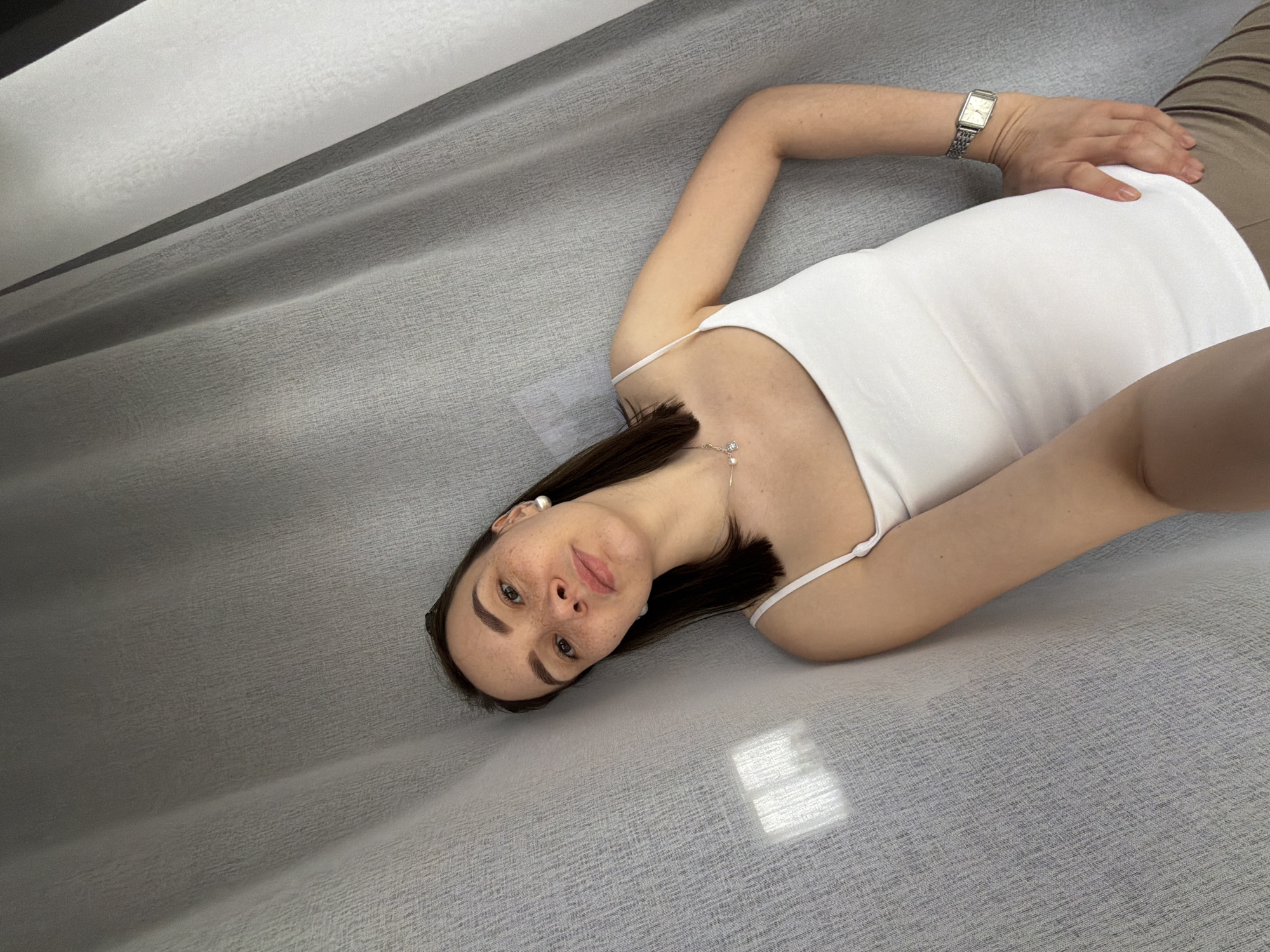A woman is lying on a gray surface. She is wearing a white tank top, brown pants, and a silver watch. She has brown hair and fair skin, with freckles on her face. She is looking at the camera. The location appears to be indoors, possibly a bed or a sofa. Light is coming through a window, creating a rectangular patch of light on the surface. Based on prior knowledge, I cannot determine the location to be Chaikovsky, Russia.