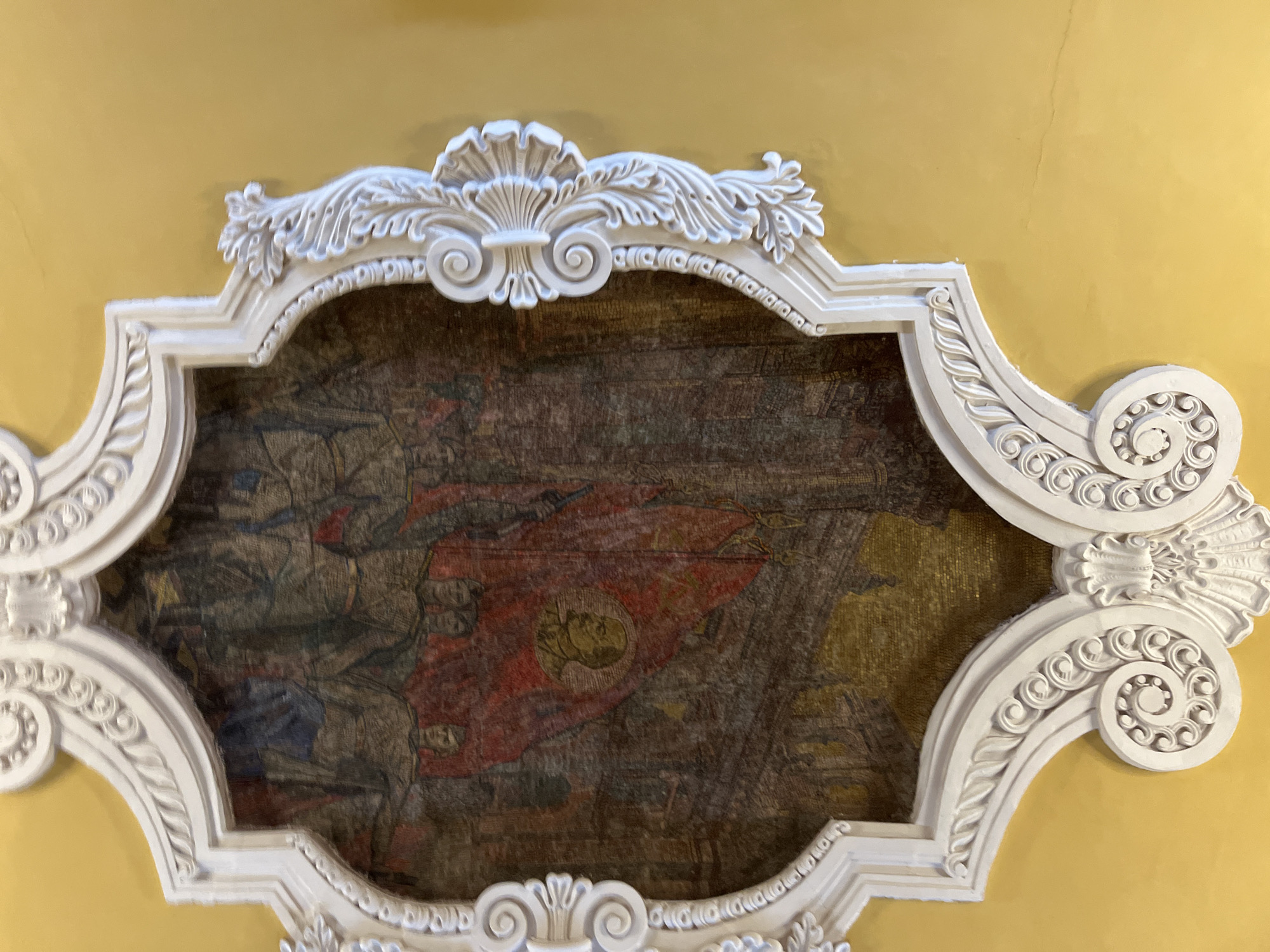 The image shows a framed mosaic or painting mounted on a yellow wall, likely within a building. The frame is ornate and white, featuring a shell-like design at the top and scrolling motifs along the sides. The artwork inside the frame appears to depict a historical or symbolic scene. A large red banner with a central emblem that includes a person's face prominently displayed. Figures and other objects in the background are unclear, suggesting a battle or gathering of people. This is likely a piece of art located in one of the stations of the Moscow Metro in Russia, judging by the style of the mosaic.