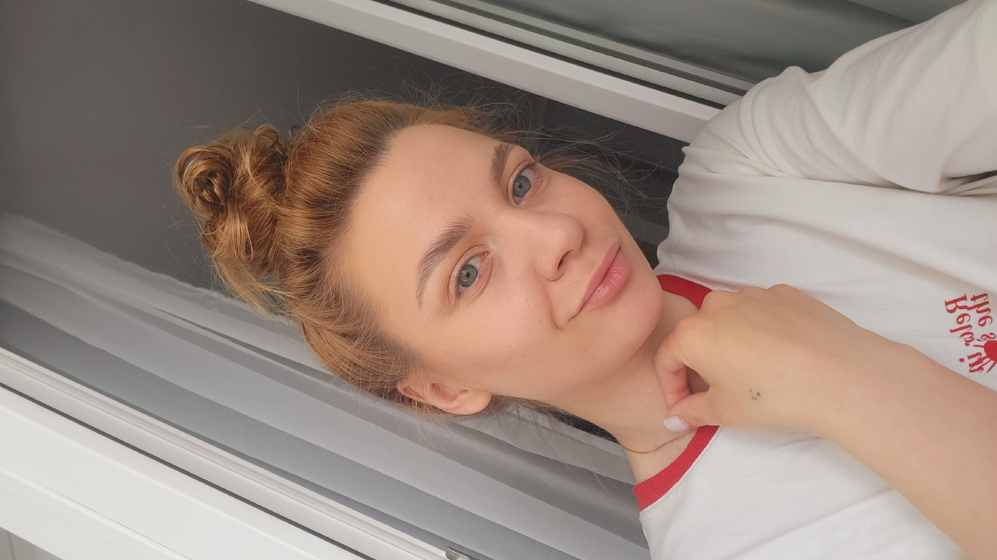 A woman is visible in a selfie-style image. She has light skin, blonde hair styled in a bun, and light blue eyes. She is wearing a white t-shirt with red trim. She is lightly touching her neck with her hand. The background shows a window with closed blinds. The image was likely taken indoors in a residential or similar setting. It is not possible to confirm the exact location of Chaikovsky, Russia from this image alone.