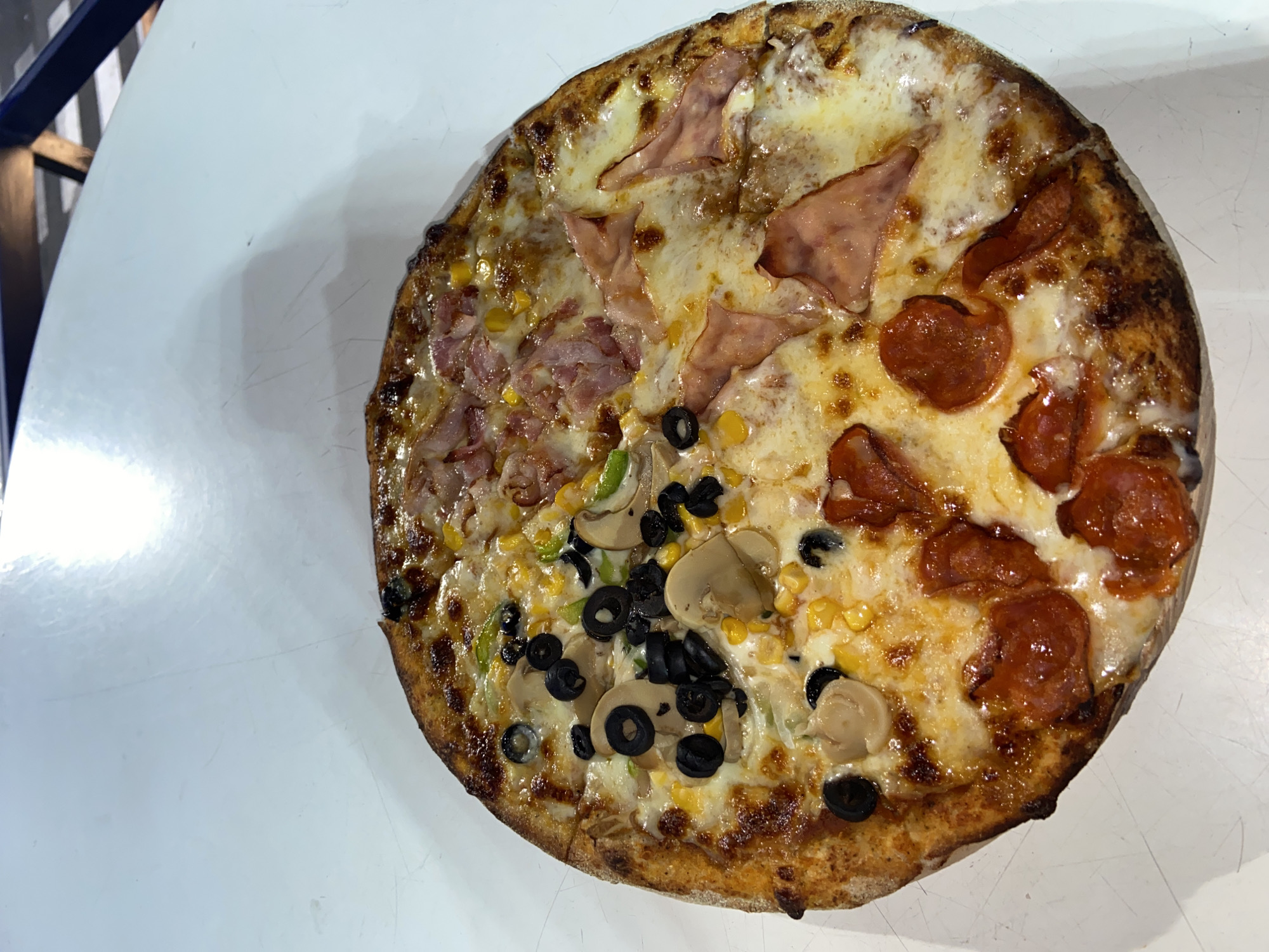 A large, round pizza with four distinct sections of toppings rests on a plain white table, likely in an eatery in Juan Griego, Venezuela. The pizza is generously covered with melted cheese and has a golden-brown, slightly charred crust.
The top-left quarter features small pieces of ham or bacon mixed with corn kernels. The top-right quarter displays larger, folded slices of ham. The bottom-left section is topped with sliced mushrooms, black olives, corn, and possibly some green peppers. The bottom-right section is covered with round slices of pepperoni or salami, along with some corn and black olives.
The table surface is bright white, showing subtle reflections and what appear to be faint scratches or marks, suggesting a used surface. In the top-left background, parts of a dark blue object and a striped pattern are visible, possibly indicating seating or wall decor. The lighting is bright and even, highlighting the textures and colors of the pizza. No people or text are visible in the image.