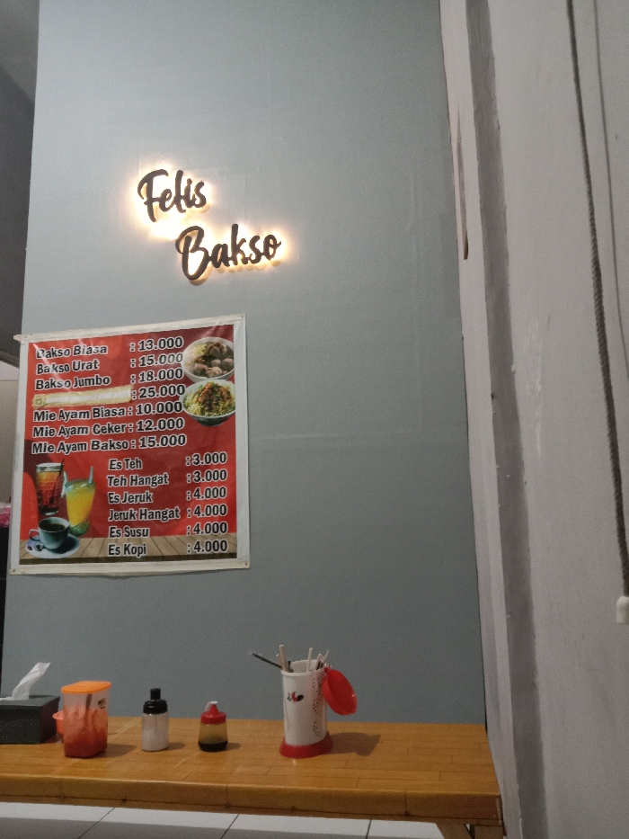 The image shows a restaurant in Kecamatan Brangsong, Indonesia, located at latitude: -6.944, longitude: 110.221. The restaurant is named "Felis Bakso" and features a bright red and white signboard with a price list for bakso (meatball soup), mie ayam (chicken noodle soup), and various drinks. The signboard is mounted on a pale grey wall. To the right of the signboard is a small table with a red plastic container of chopsticks, a white bottle with a red cap, a red bottle with a white cap, and a red container with a white lid. The table is wooden and has a light brown finish. There is a white wall to the right of the table. The environment is clean and well-lit.