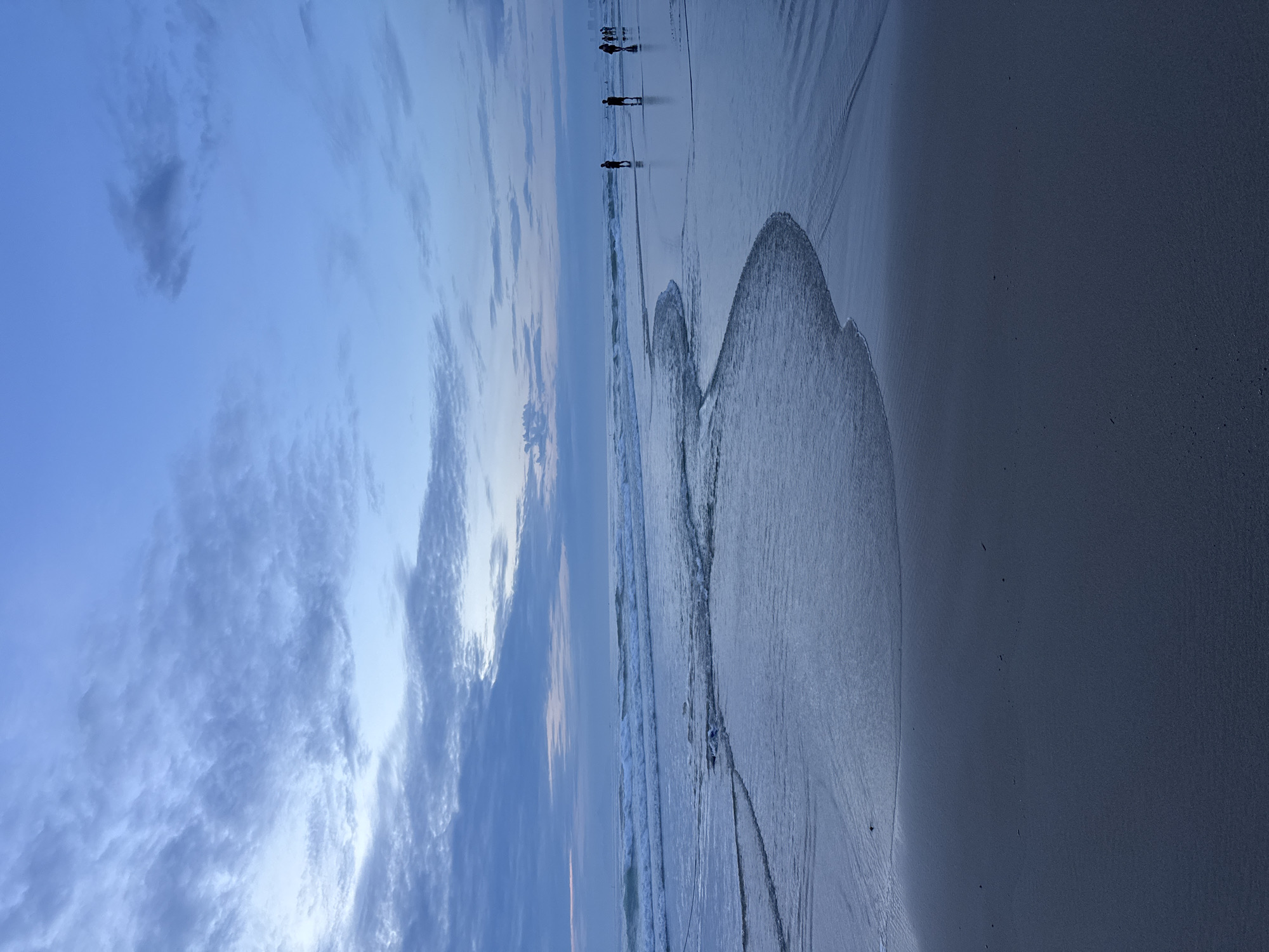 The image shows a beach at dusk or dawn. The sky is blue with some clouds. The beach is sandy and wet, with some waves coming in. There are three people walking along the beach in the distance.
Without additional information, determining a specific location within the city of Đà Nẵng is not possible.