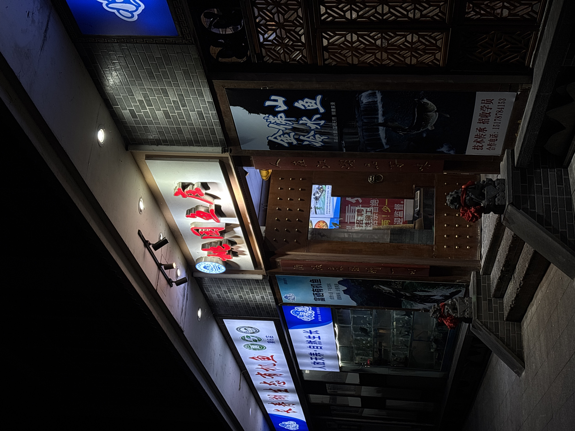 The image is taken at night and shows an angled view of storefront signs and building elements. The location appears to be in Nan'an District, Chongqing, China based on the information on the signs.
Visible signage includes:
- Signs advertising "富硒有机鱼" (Selenium-rich organic fish) and "金佛山 乐鱼" (Jinfo Mountain Leyu Fish), suggesting a restaurant specializing in fish dishes.
- A recruitment notice for a dishwasher.
- A sign mentioning "技术传承 招收学员" (technical inheritance, recruiting students) and providing a phone number.
Architectural details include brickwork, decorative woodwork, and illuminated signs. The overall environment is dark, with the signs providing the primary source of light. There are stairs and elements suggesting traditional Chinese architectural design.