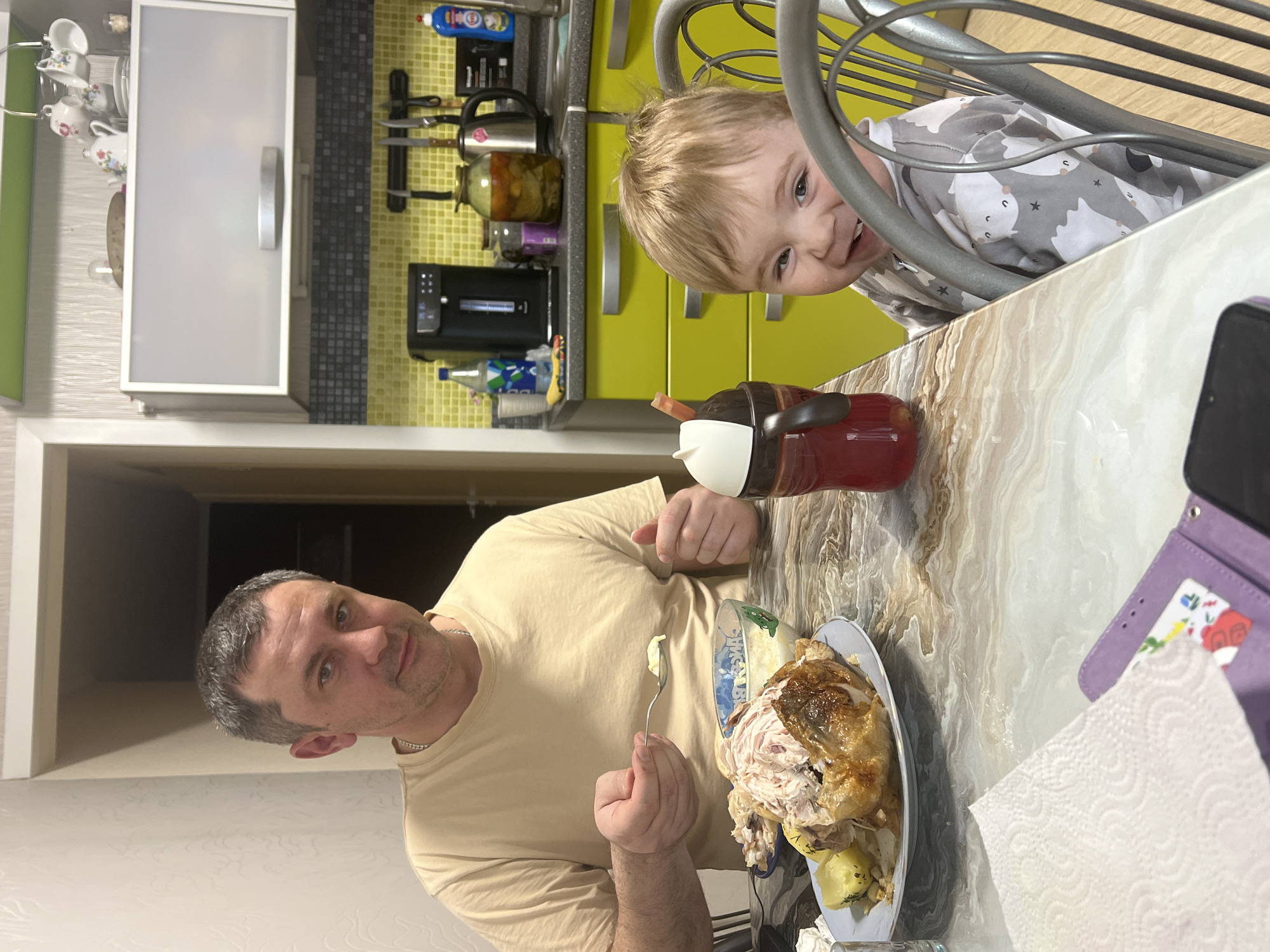 The image captures an indoor scene, likely a kitchen and dining area in a home in Perm, Russia. The perspective is tilted, showing a man and a young child engaged at a dining table.
**Subjects:**
A man with short, dark gray hair and a light-colored (beige) t-shirt is seated at a table, looking directly at the camera. He is holding a fork with a small amount of food on it, suggesting he is in the middle of a meal.
A young child with blond hair is playfully peeking over the table from behind a metal chair. The child wears a grey long-sleeved shirt patterned with white cats/foxes and stars, and has a wide, joyful smile, looking towards the camera.
**Objects:**
The dining table has a light-colored, marble-like pattern. On the table are a large plate with a partially eaten roasted chicken and pieces of potato, a bowl of white salad (possibly coleslaw), and a red beverage bottle with a white and black lid. To the right, a purple wallet or organizer with a colorful card and a white paper napkin are visible.
In the background, a kitchen counter features bright lime green lower cabinets with metallic handles and gray upper cabinets with frosted glass doors and metallic handles. The backsplash is a gray and black mosaic tile pattern. Various kitchen items are on the counter, including a silver kettle, several glass jars (one with preserves), a black coffee machine, and a blue bottle of cleaning product on a rack.
**Activity or event taking place:**
The man is eating a meal, possibly dinner, while the child playfully interacts with the scene, perhaps peeking at the adult or for attention from the person taking the photo. It appears to be a casual family mealtime.
**Notable details:**
The indoor setting, illuminated by artificial lighting, suggests the time of day could be evening or night. The child's expression is one of clear joy and playfulness, with a wide, open-mouthed smile. The man's expression is neutral to slightly pleased, as he looks at the camera. The kitchen design, including the type of cabinets and jars on the counter, offers some subtle cultural cues consistent with a home in Russia.
**Visible text:**
On the blue bottle of cleaning product in the background, the word "Whirlpool" is visible in English, along with some smaller, illegible Cyrillic text.
