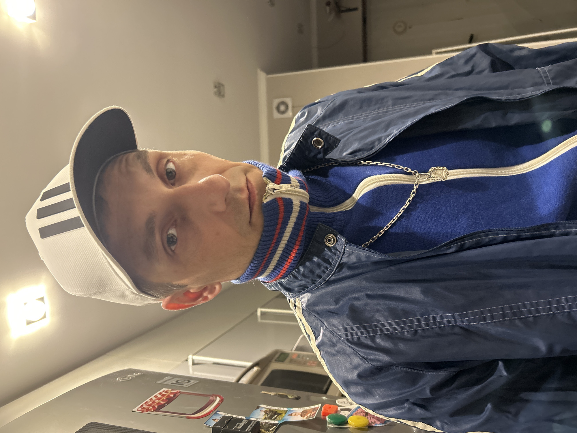 A man in his 30s is posing for a selfie in what appears to be a kitchen in Pushkino, Russia. He is wearing a white baseball cap with black stripes, a blue turtleneck sweater with white and red stripes around the collar, and a navy blue track jacket with white stripes on the sleeves. He has a silver chain with a pendant around his neck. The background shows a refrigerator with several magnets and stickers, kitchen cabinets, and a light fixture on the ceiling. The overall impression is casual and informal.