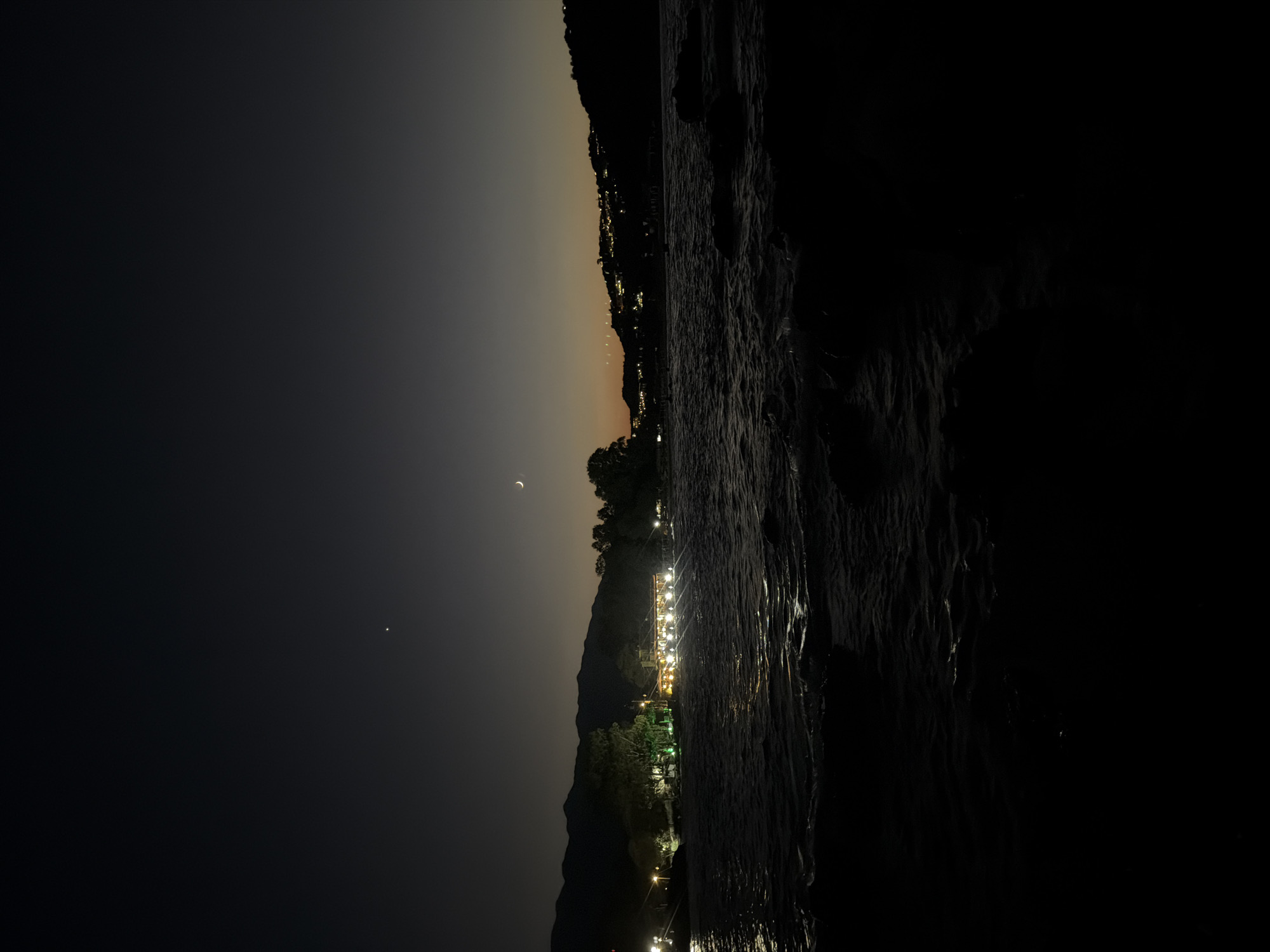 The image depicts a nighttime scene along the coast of Georgioupoli, Greece. The foreground is dominated by dark, choppy water, with some rocks visible at the very bottom. Across the water, a well-lit building with outdoor seating, possibly a restaurant or taverna, is illuminated by numerous yellow lights. Trees surround the building, and further in the background, rolling hills are visible. Scattered lights indicate buildings on the hillsides, suggesting a town or village. The sky is a dark twilight hue, with a faint crescent moon and a bright star visible in the upper portion. The overall atmosphere is tranquil and serene. There are no people visible in the image.