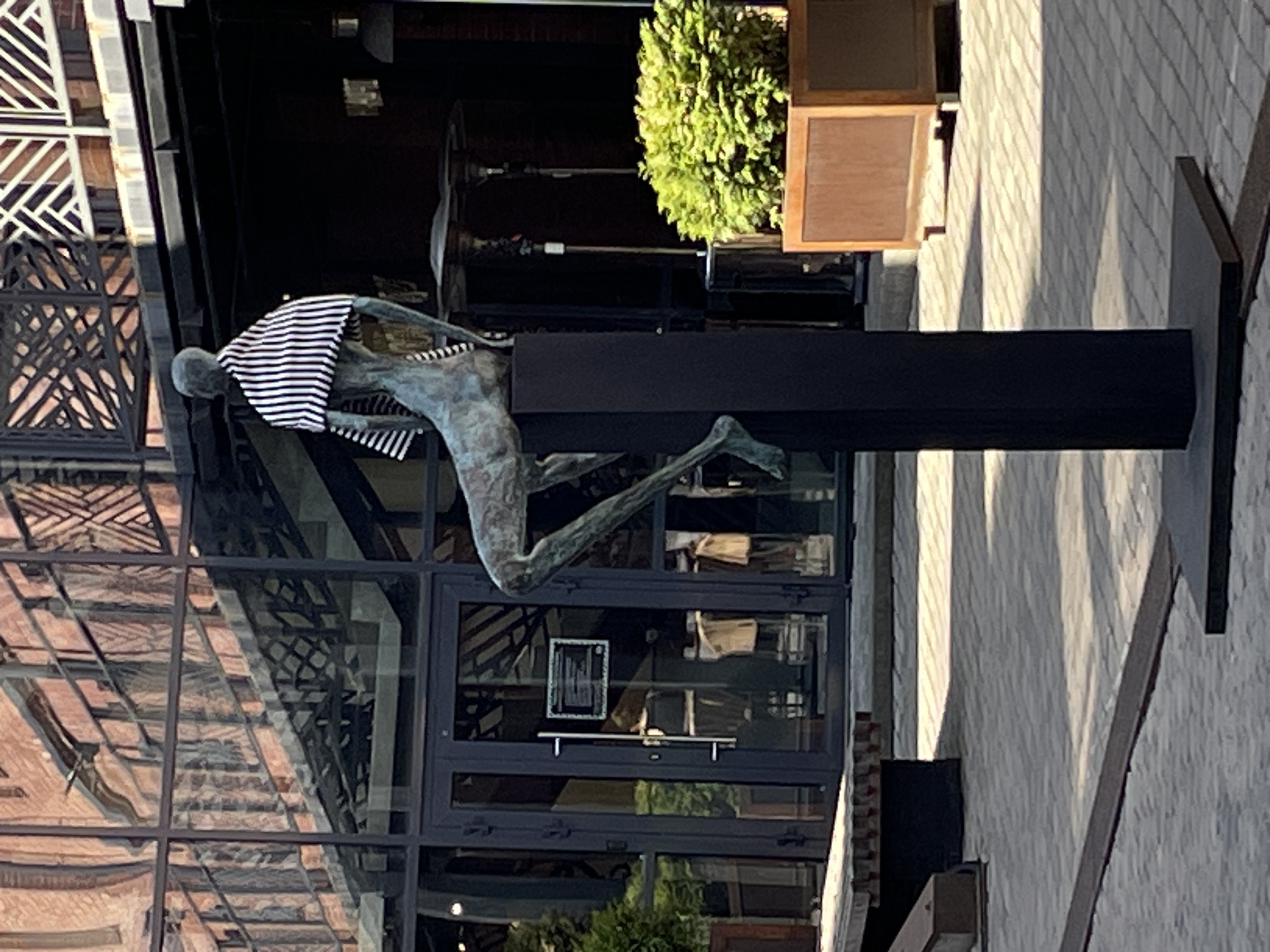 A statue of a man in a striped shirt and running posture is placed on a black rectangular stand. The statue is situated in front of a building with glass windows and a reflection of brick structures. A bush is in a wooden planter to the right of the statue. The statue and building are located on a paved area. Based on architectural style and urban characteristics, the statue is likely in Saint Petersburg, Russia.