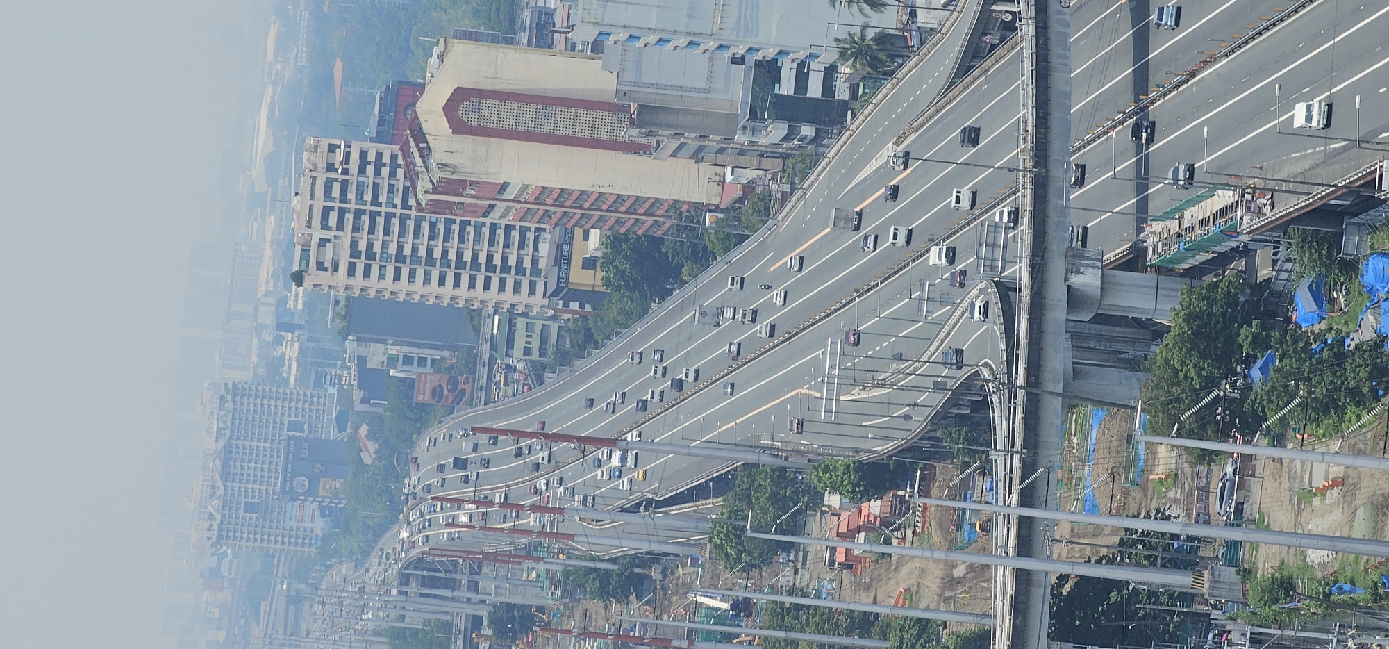This aerial photograph depicts a multi-lane elevated highway, characteristic of the city of Makati, Philippines. The highway is densely populated with vehicles, including cars, vans, and trucks, suggesting moderate traffic flow. On either side of the highway, numerous high-rise buildings, typical of Makati's urban landscape, dominate the skyline. Some of these buildings appear to be commercial establishments, with signage visible on their facades. Construction activity is also evident in certain areas, indicated by scaffolding, building materials, and heavy machinery, which further points to ongoing development in the city. The sky is hazy, a common atmospheric condition in urban environments, partially obscuring the distant structures. The perspective suggests the photograph was taken from a very high vantage point, likely a tall building or a drone.