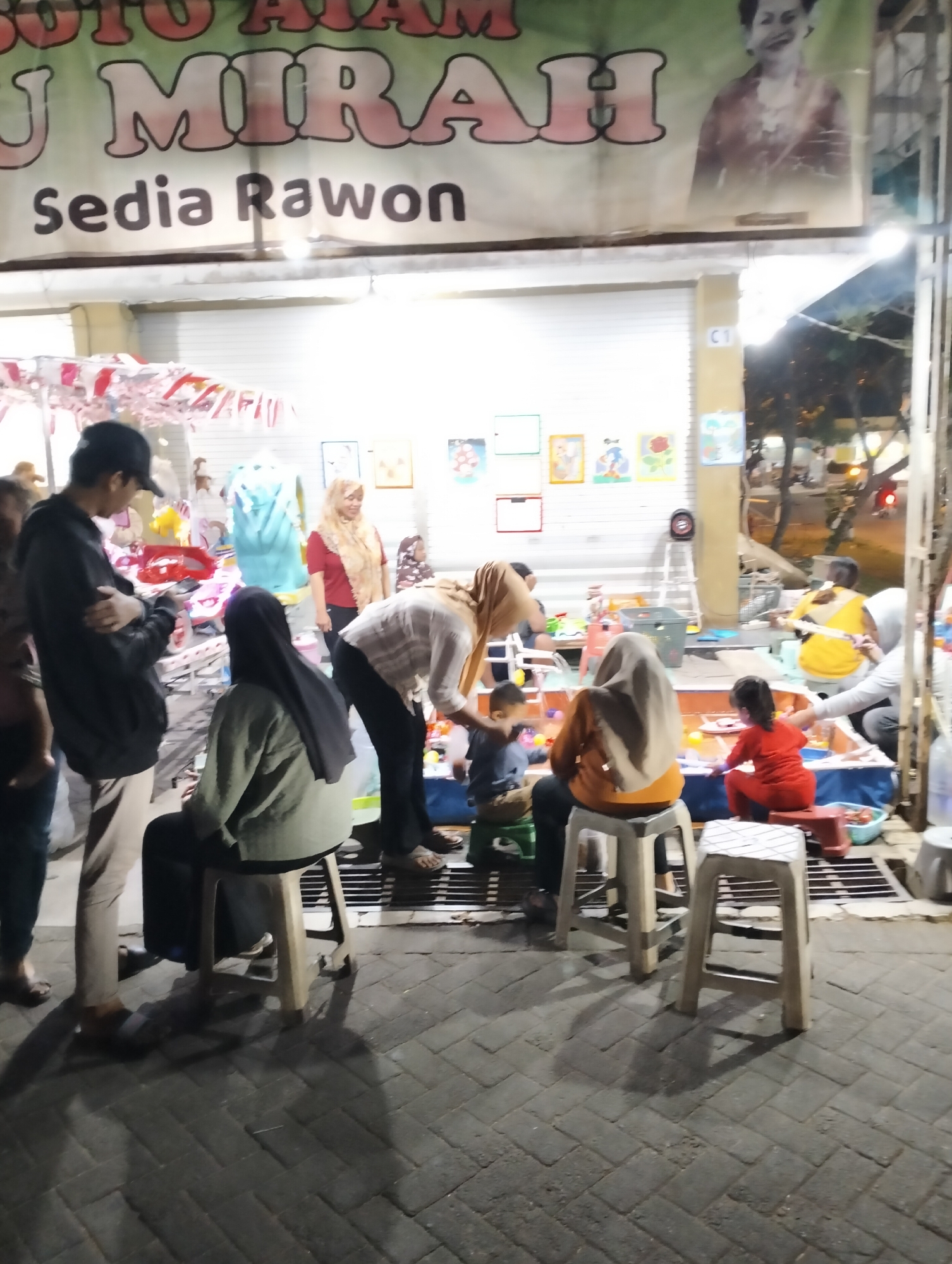 This image captures an outdoor scene at night, likely a food stall or small eatery in Semarang, Indonesia, as suggested by the Indonesian text on the banner. The banner reads "SOTO AYAM MIRA" and "Sedia Rawon," indicating that the establishment serves Soto Ayam and Rawon, popular Indonesian dishes.
In the foreground, several people are visible. A man wearing a black hoodie and a black baseball cap is standing, looking at his phone. To his left, another person's legs are visible. In the center, a woman wearing a black headscarf and a light green jacket is sitting on a stool, facing away from the camera. Next to her, a woman wearing an orange headscarf is also sitting on a stool, facing towards a shallow water-filled area. A young child in an orange outfit is seated in front of the water, interacting with colorful balls. Another woman, wearing a white and brown patterned shirt and a hijab, is bending over the water area.
Further back, several other individuals are seen. A woman in a red shirt and a beige headscarf is standing and appears to be engaged in conversation or serving. A young child is also visible near the water. On the right side of the image, a person in a yellow shirt is seated, and other figures are in the background, some of whom are patrons of the establishment.
The environment appears to be paved with brick or stone tiles. Various items are present, including stools, tables, and what looks like play or activity stations, possibly for children. The overall lighting suggests it is evening or night, with artificial lights illuminating the area. The scene conveys a casual, community-oriented atmosphere typical of street food vendors or small local businesses.