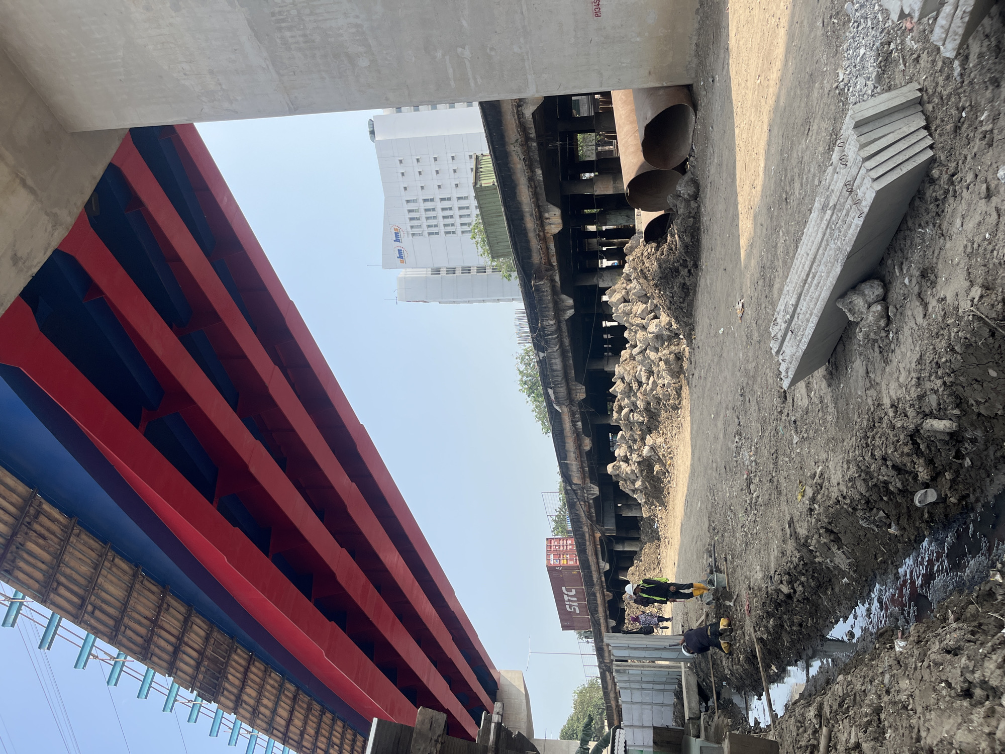 In North Jakarta, Indonesia, a construction site beneath an elevated red and blue bridge is depicted. The scene is dominated by the unfinished infrastructure, with the large red beams of the bridge structure visible overhead. The ground is a mix of dirt and debris, with piles of rocks and large metal pipes scattered around.
Two construction workers are present. One, wearing a yellow hard hat and a reflective vest, is standing near a trench filled with murky water. Another worker, also in a hard hat and reflective vest, is kneeling by the trench, possibly engaged in some form of manual labor. Several other individuals are walking in the background, near a shipping container on an elevated structure.
In the distance, a modern white building with the logo "ubm bms" is visible against a clear blue sky. The overall impression is one of ongoing development and construction in an urban environment.