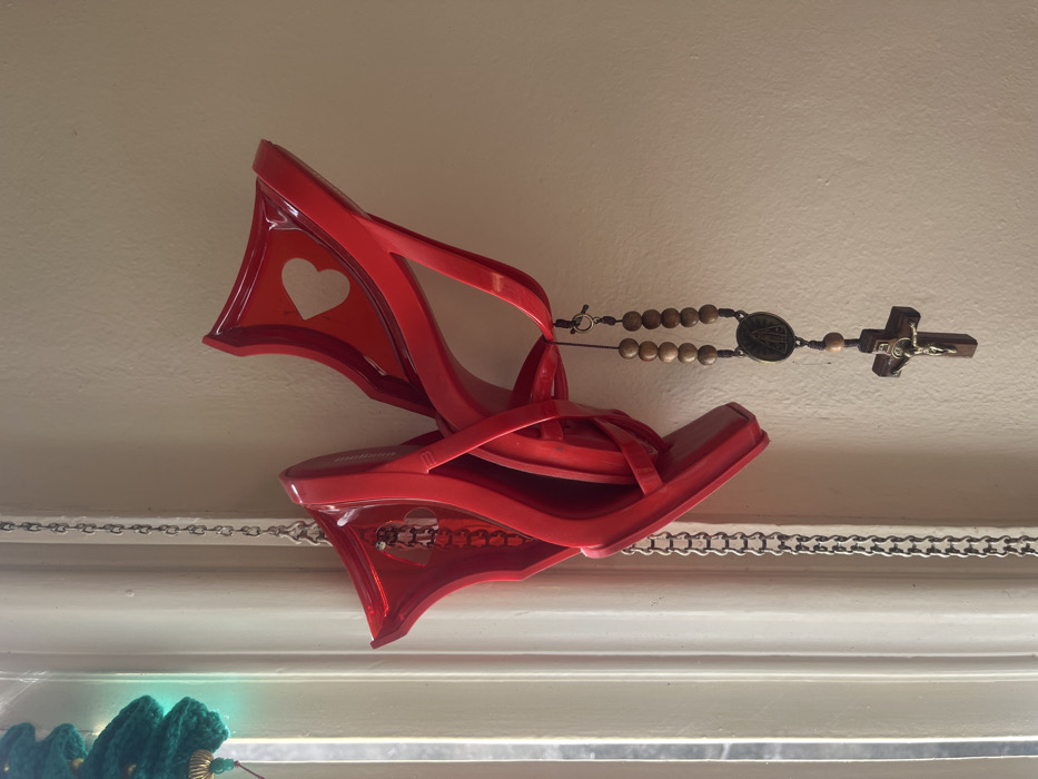 The image displays a pair of bright red, high-heeled shoes hanging indoors, suspended by a metallic chain visible on the left. The shoes, appearing to be made of a glossy synthetic material, are interlocked, with the shoe on the upper right featuring a heart-shaped cutout on its upper portion. A rosary, composed of light-colored wooden beads, a dark cord, a religious medallion depicting a figure, and a wooden crucifix with a metal corpus, is suspended from one of the red shoes. The background consists of a light-colored wall on the right and a white-painted window frame or trim on the left, suggesting an indoor setting. A blurred green knitted or crocheted object is partially visible in the upper-left corner near the window. Natural light illuminates the scene from the left. The specific location within Buenos Aires, Argentina, cannot be determined from the visual information provided.