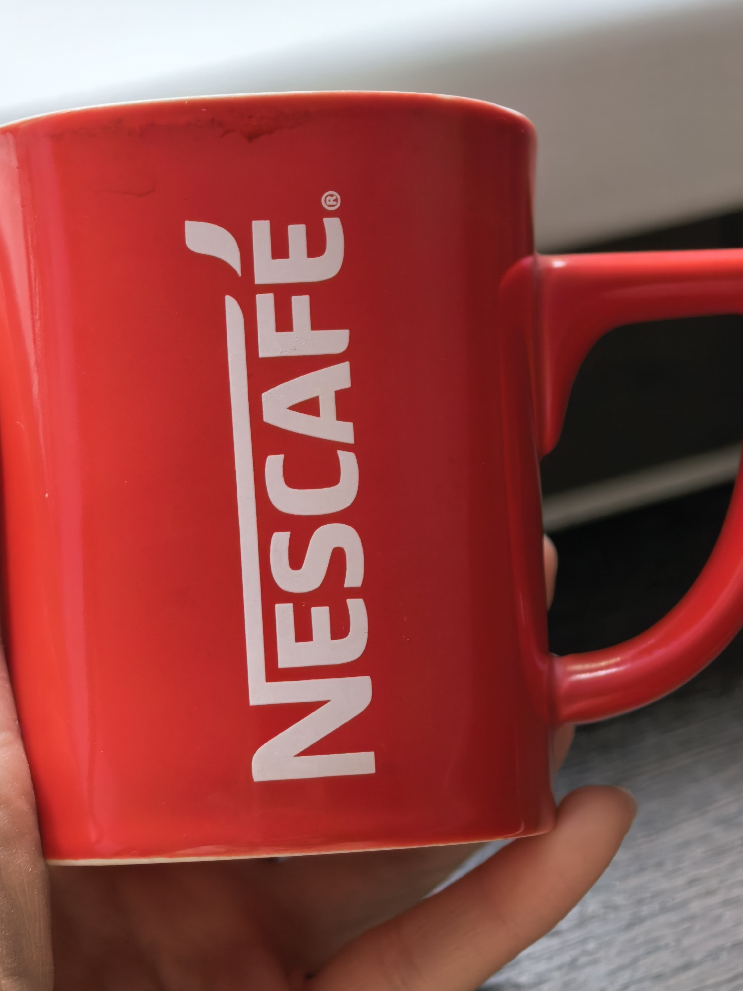 A close-up photograph shows a red mug with the word "NESCAFÉ" printed vertically in white. The mug is being held by a hand. The background is a mix of white and dark gray tones. The location cannot be determined from the image.