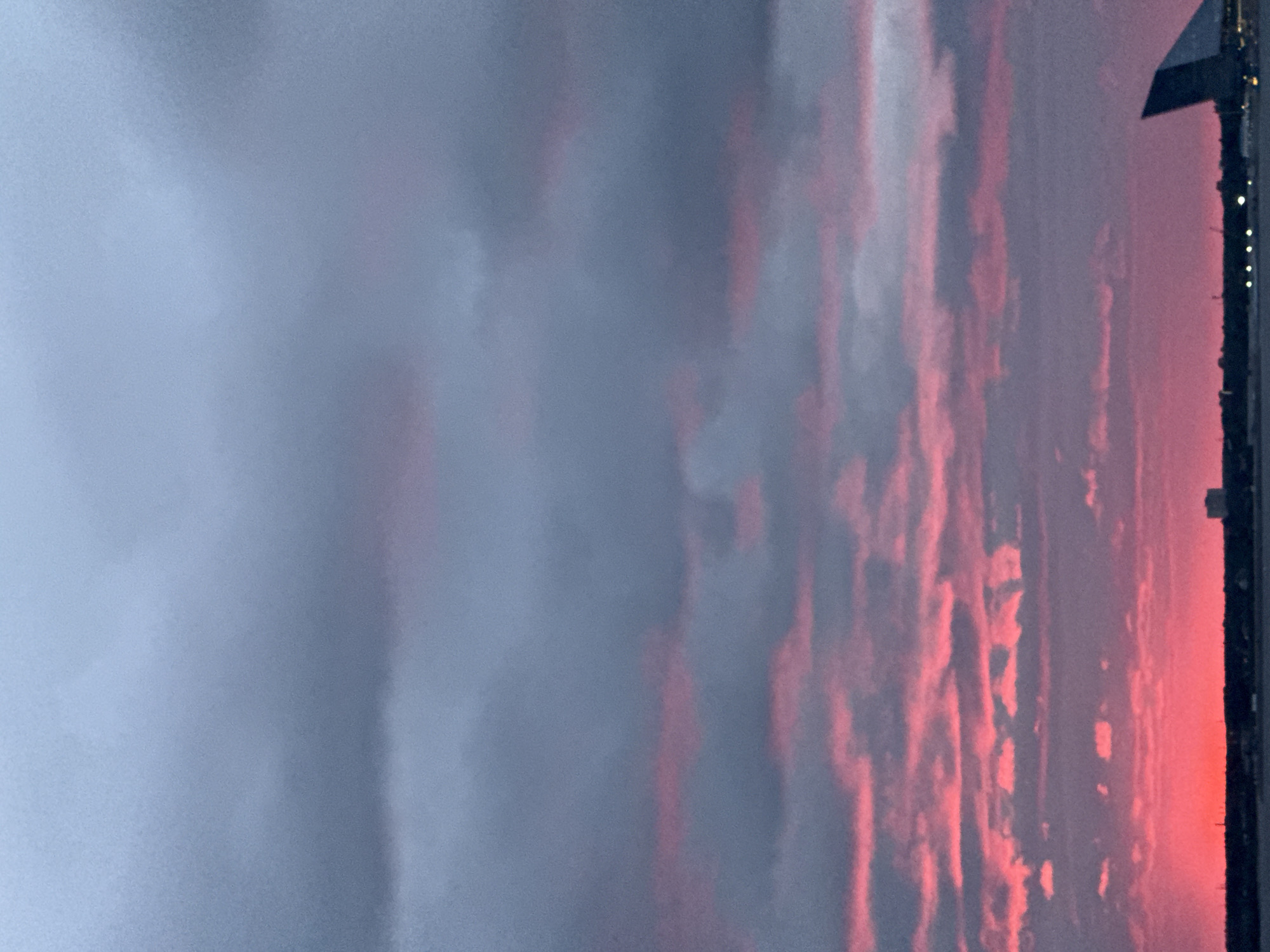 The image captures a twilight sky over a body of water, likely the Gulf of Finland near Saint Petersburg, Russia. The sky is dominated by dramatic clouds, with streaks of vibrant pink and red illuminating the lower portion, indicative of a sunset or sunrise. Above these colorful clouds, the sky transitions to shades of gray and blue, with darker, more ominous clouds forming the upper layer.
Along the horizon, a dark silhouette of a city skyline is visible, featuring buildings of varying heights. Among these structures, a distinct, modern building with a pointed, angular roof is prominent on the right side of the frame, possibly part of the St. Petersburg Stadium or a related architectural complex. Lights from the city twinkle along the waterfront, suggesting it is evening. The water surface is dark and still, reflecting the dim light of the sky. There are no people or active subjects discernible in the image. The overall impression is one of a moody and atmospheric celestial display over an urban coastal landscape.