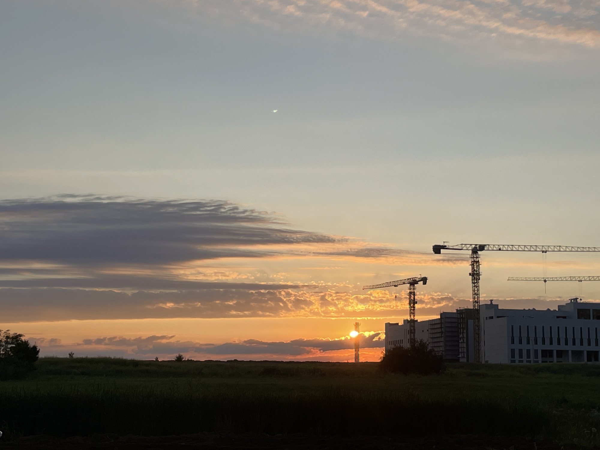 The image depicts a sunset scene with construction cranes and a modern building under construction. The sky is filled with a gradient of colors from light blue at the top to warm orange and pink hues near the horizon, indicating the sun is setting. Several clouds are visible, some wispy and others more defined, catching the colors of the sunset. In the upper part of the sky, a sliver of a crescent moon is visible.
In the middle ground, a silhouette of a grassy field is present. On the right side of the image, a large, modern white building with many windows is partially constructed. Several tall construction cranes are positioned around the building and in front of it, suggesting ongoing development. The sun appears to be setting behind one of the cranes, creating a bright glow. The foreground consists of a dark silhouette of vegetation, possibly grass or bushes.
Without specific geographical markers in the image, it is impossible to definitively identify the location as Stavropol, Russia. The scene is characteristic of a construction site on the outskirts of a city or in a developing area. The time of day is clearly sunset due to the lighting and sky colors. There are no people visible in the image. The overall mood is one of development and the transition from day to night.