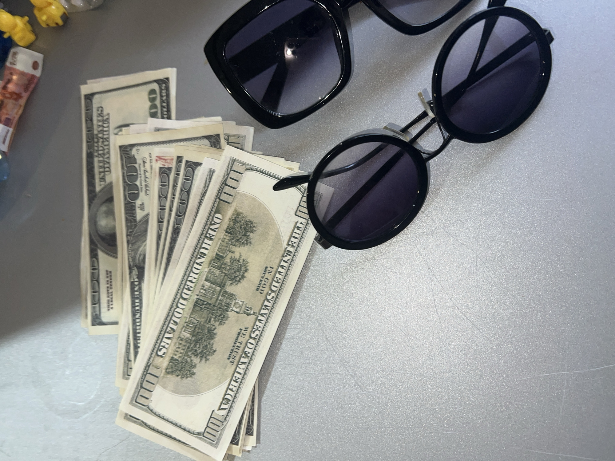 The scene displays a collection of items arranged on a light gray, textured surface, possibly a tabletop, exhibiting multiple scratches. In the upper left, a fanned stack of approximately ten $100 U.S. dollar bills is visible. Closer examination of these bills indicates they are explicitly marked "THIS NOTE IS NOT A LEGAL TENDER" and bear "SOUVENIR" or "FOR COLLECTION PRODUCTION" text, identifying them as prop or novelty currency. To the right, two pairs of sunglasses are present. The upper pair features large, dark, square-framed lenses, while the lower pair consists of smaller, round black frames with dark lenses. The upper right background shows blurred objects, including what appears to be a rolled or folded European banknote, possibly a Euro, and several small, yellow, plastic items. No individuals are depicted. The precise location within Noviy, Russia, cannot be determined from the visual information provided.