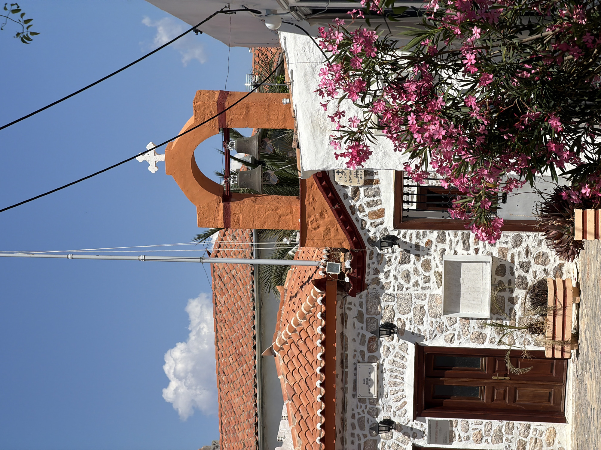 The image depicts a building in Hydra, Greece, characterized by its stone masonry construction and tiled roof. A prominent orange-colored bell tower with three bells is visible, topped with a white cross. The building's exterior features a wooden door, a window with wooden shutters, and flowering shrubs. Power lines run across the top of the frame. The sky is partially visible with some clouds.