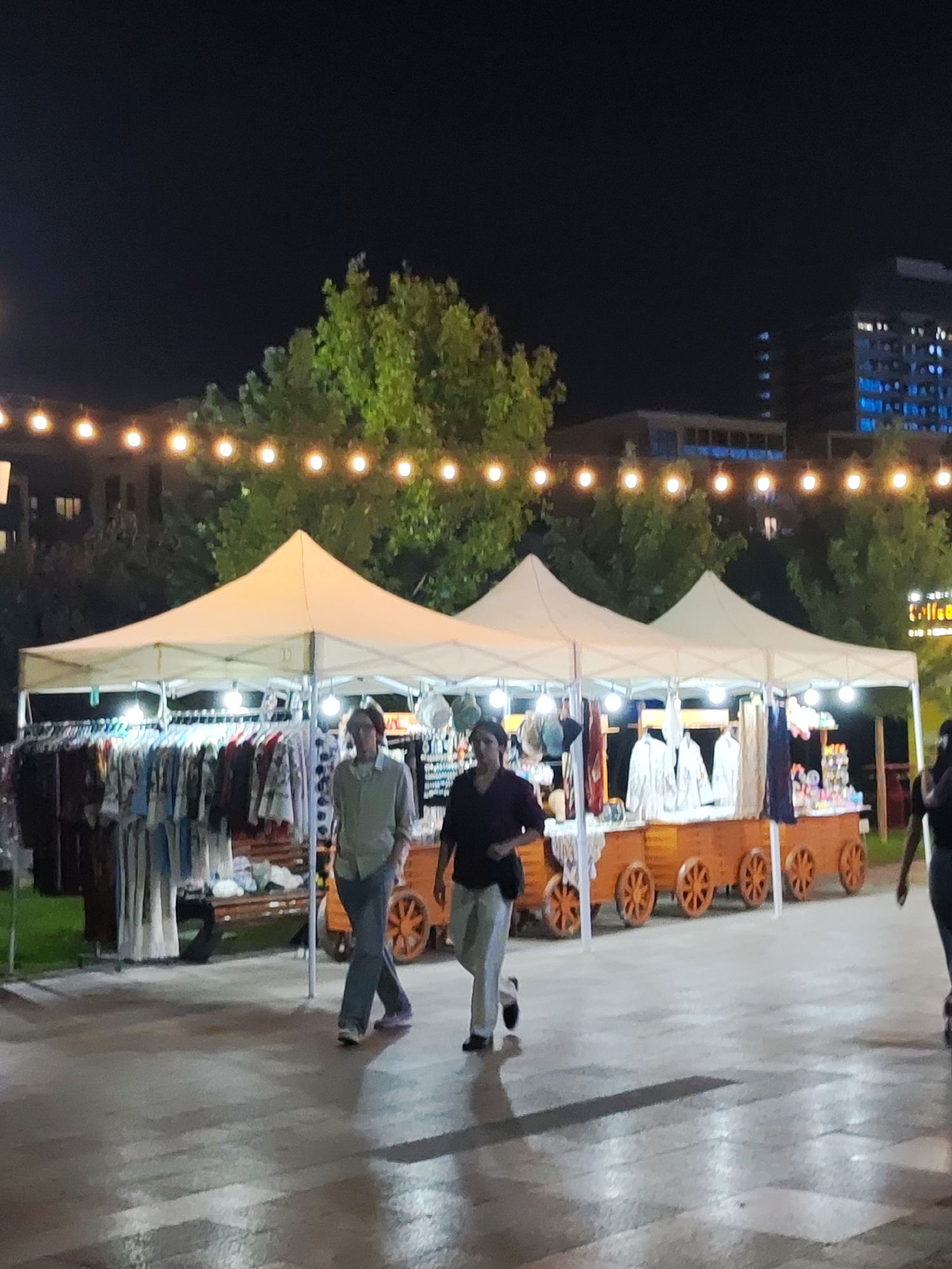 The image depicts an outdoor market scene at night in Tashkent, Uzbekistan. Two white pop-up tents are set up, displaying various clothing items on racks and shelves. The tents are illuminated by string lights overhead and internal lighting. In the foreground, two individuals are walking across a paved area. The person on the left is wearing a light-colored shirt and jeans, while the person on the right is wearing a dark top and light-colored pants. Behind the tents, trees and modern buildings are visible, with one building displaying illuminated blue windows. The market appears to be set up in a public square or walkway.