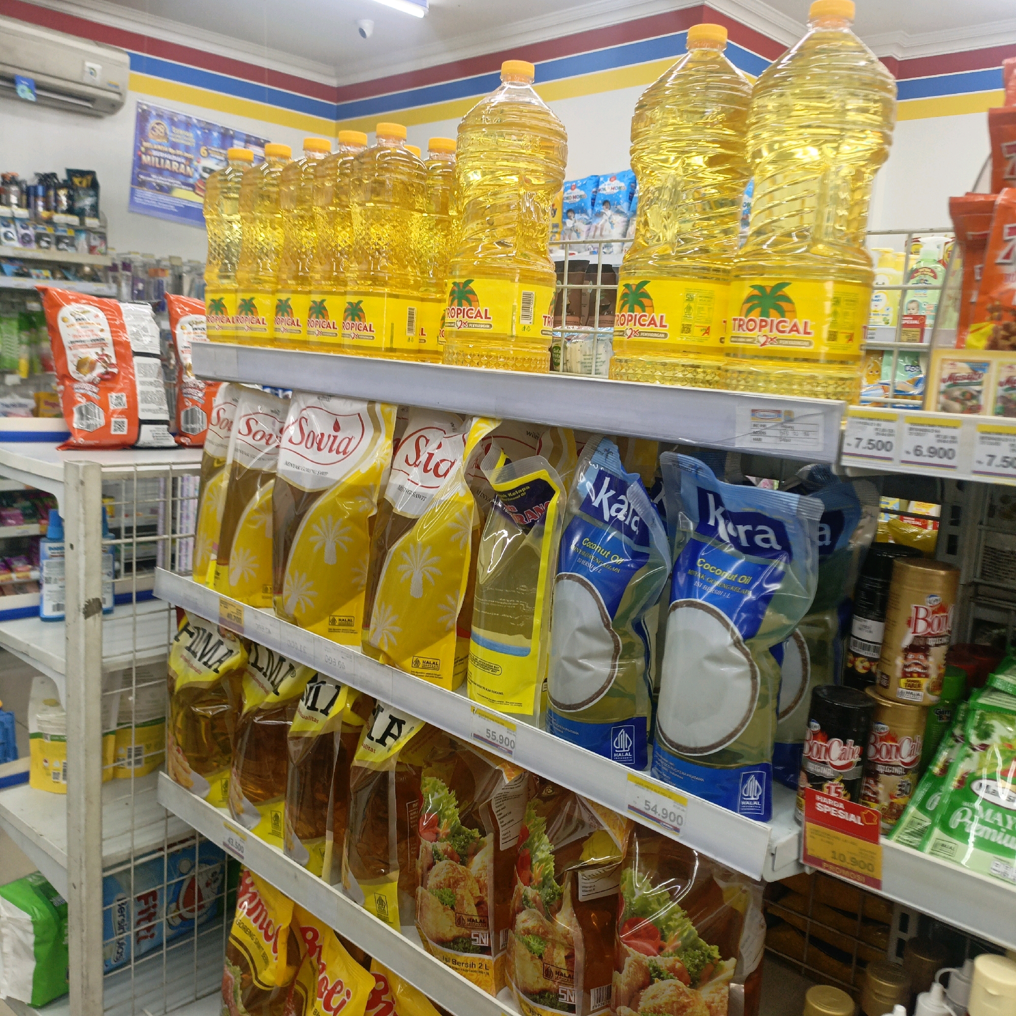 The image depicts a well-stocked retail environment, likely a minimarket or convenience store, with shelves filled with various consumer goods. The dominant products are cooking oils in large plastic bottles and pouches, prominently featuring "Tropical" branding with palm tree graphics. There are also pouches of "Sovia" and "Sia" cooking oil, and "Kara" coconut oil. Other items visible include bagged snacks, canned goods labeled "Bon Cade," and packets of "Maya" mayonnaise. The shelving is typical of retail displays, with price tags clearly visible on some items. The store appears clean and organized. The presence of Indonesian text on the product packaging, along with the "Halal" certification, suggests the location is in Indonesia. Given the specific arrangement of the store and its branding, this scene is consistent with a minimarket in Salatiga, Indonesia. There are no people visible in the image.