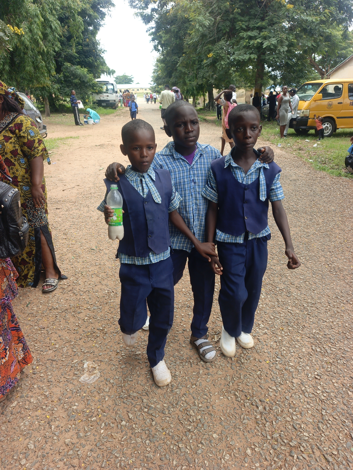 The image depicts a group of people on a gravel path, likely in Akwanga, Nigeria. In the foreground, three young boys are walking, dressed in school uniforms. The boy on the left wears a blue and white checkered shirt with a dark blue vest and pants. The middle boy is similarly dressed, holding a bottle of liquid in his right hand. The boy on the right, also in uniform, has his arm around the middle boy. Behind them, a woman in a vibrantly patterned dress is visible, carrying a dark bag. Further in the background, various other individuals are present, including adults and children, some near a yellow bus and a white van. The environment is characterized by lush green trees lining the path, suggesting a rural or semi-rural setting within Akwanga. The ground is covered with gravel and small stones, with some scattered yellow petals from trees. The overall impression is of a school or community gathering outdoors.