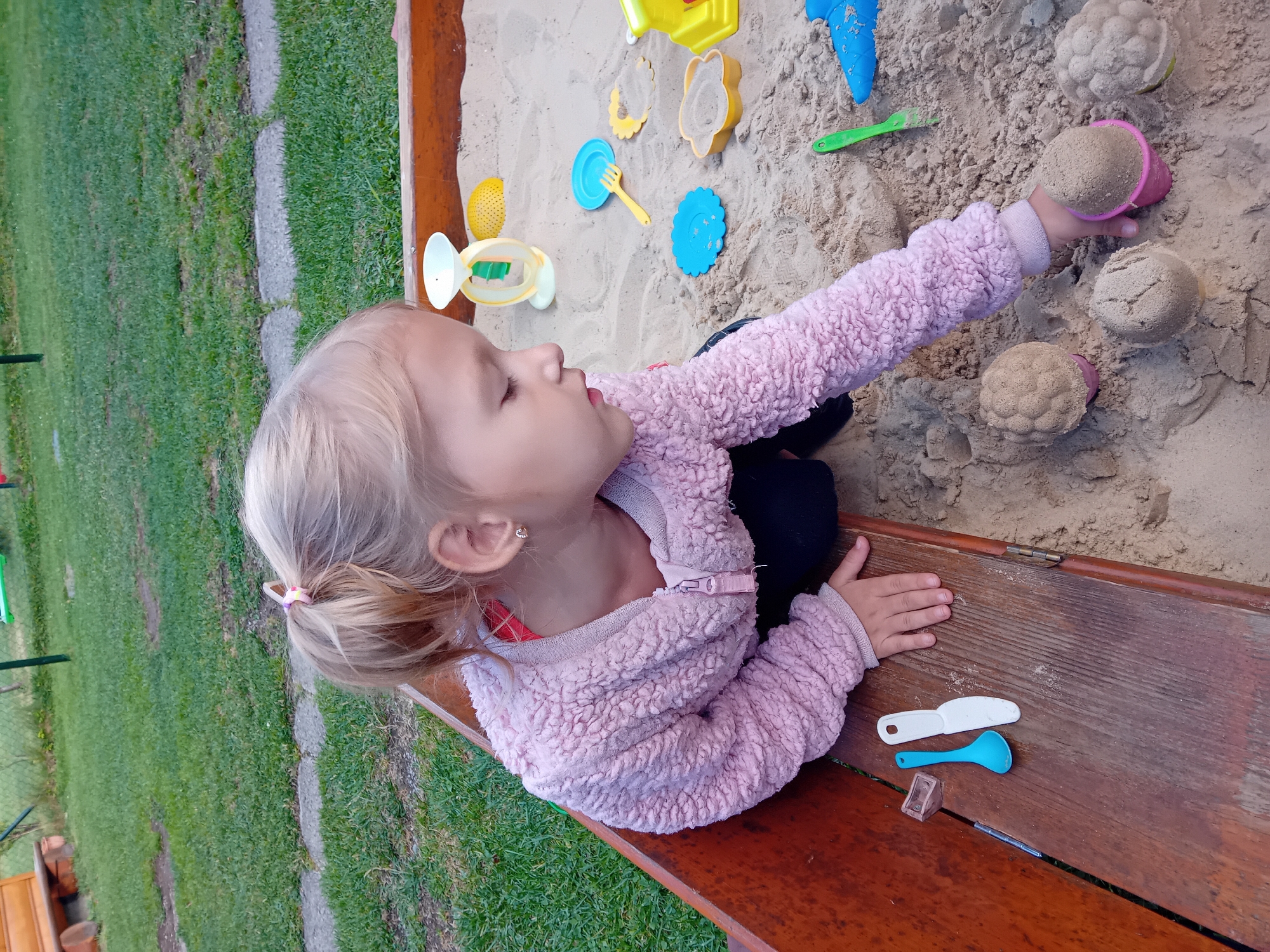 A young Caucasian girl with blonde hair tied in a ponytail is playing in a sandbox. She is wearing a pink fuzzy jacket over a red shirt and black pants. She is crouched down in the wooden sandbox, with her right hand reaching towards a pink sand mold filled with sand. There are several other sand molds, toys, and sand creations in the sandbox, including a yellow balance scale toy, blue plates and a fork, and a yellow house-shaped toy. The sandbox is located outdoors, with green grass and a wooden bench visible in the background. This scene appears to be in the city of Zuberec, Slovakia, as indicated by the presence of a traditional Slovakian wooden structure in the background, which is characteristic of the region's architecture and homesteads. The lighting suggests it is daytime, possibly late afternoon given the soft light.