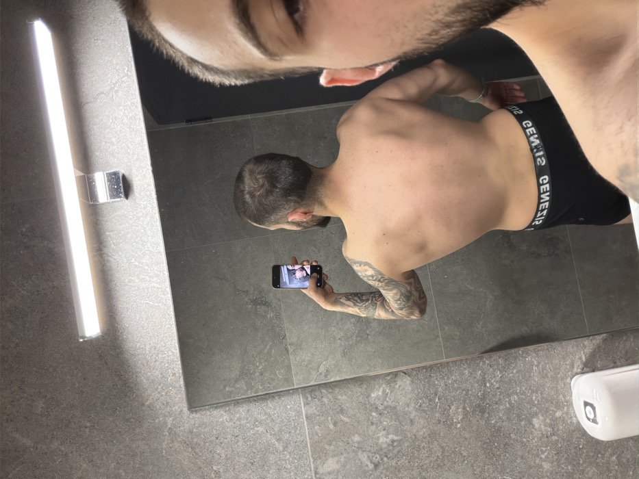 A male individual is depicted taking a mirror selfie in an indoor setting, identified as a bathroom or changing room in Plovdiv, Bulgaria. The subject is primarily visible in reflection, standing with his back partially turned and his left side exposed. He possesses a dark, patterned sleeve tattoo covering his left arm. He wears black boxer briefs featuring a white waistband with the discernible text "GENESIS" (appearing reversed as "SISENƎӘ" due to the mirror). His short, dark hair and beard are visible in the reflection, with a portion of his face and upper shoulder also present in the extreme right foreground, outside the mirror's reflection. A black smartphone is held in his left hand, oriented towards the mirror to capture the image. The immediate environment includes large, rectangular grey textured wall tiles and a horizontal LED light fixture mounted above the mirror. A white, wall-mounted dispenser is partially visible in the lower-left foreground.