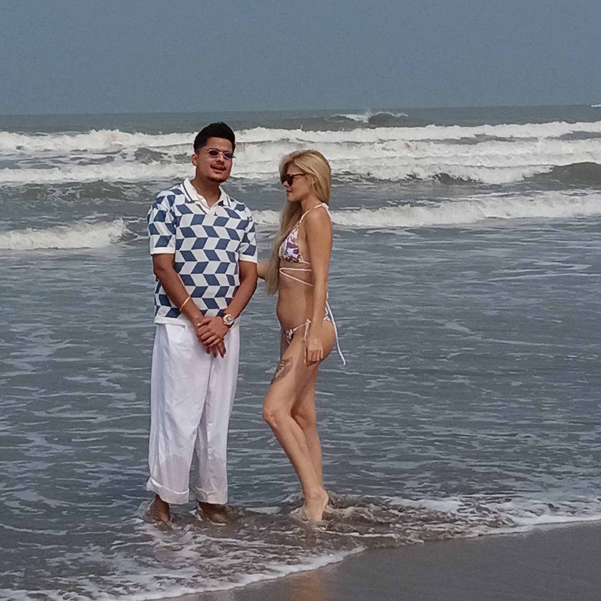 The image depicts a man and a woman standing in the water on a beach, seemingly engaged in conversation. The location is likely Mandrem, India, given the beach setting. The man is wearing a blue and white patterned short-sleeved shirt and white pants. He is also wearing sunglasses and a watch. The woman is wearing a floral bikini and sunglasses. She has a tattoo on her leg. The ocean is visible behind them with breaking waves. The sky is overcast.