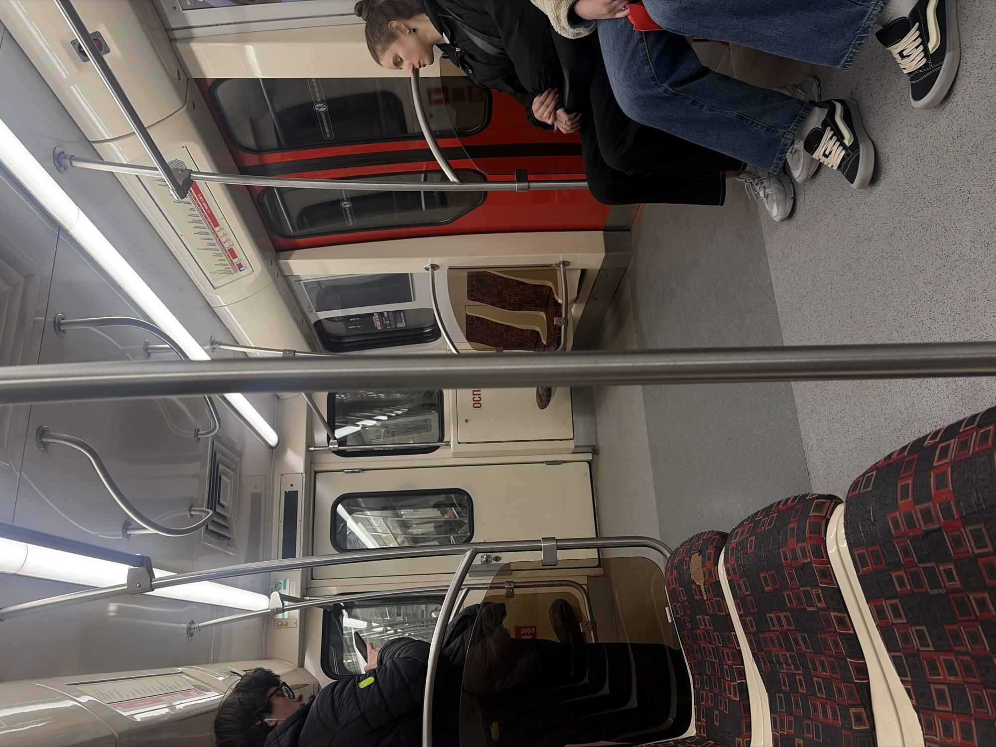 The image shows the interior of a subway car in Saint Petersburg, Russia. Two people are standing, with one facing forward and the other with their head tilted down. To the right are two sets of legs. Seated in the foreground is a person wearing a dark jacket and glasses, looking at a device in their hand. The seats have a patterned fabric cover, with red, black, and white squares. Overhead are handrails. The walls are white, and there are windows and doors along the sides.