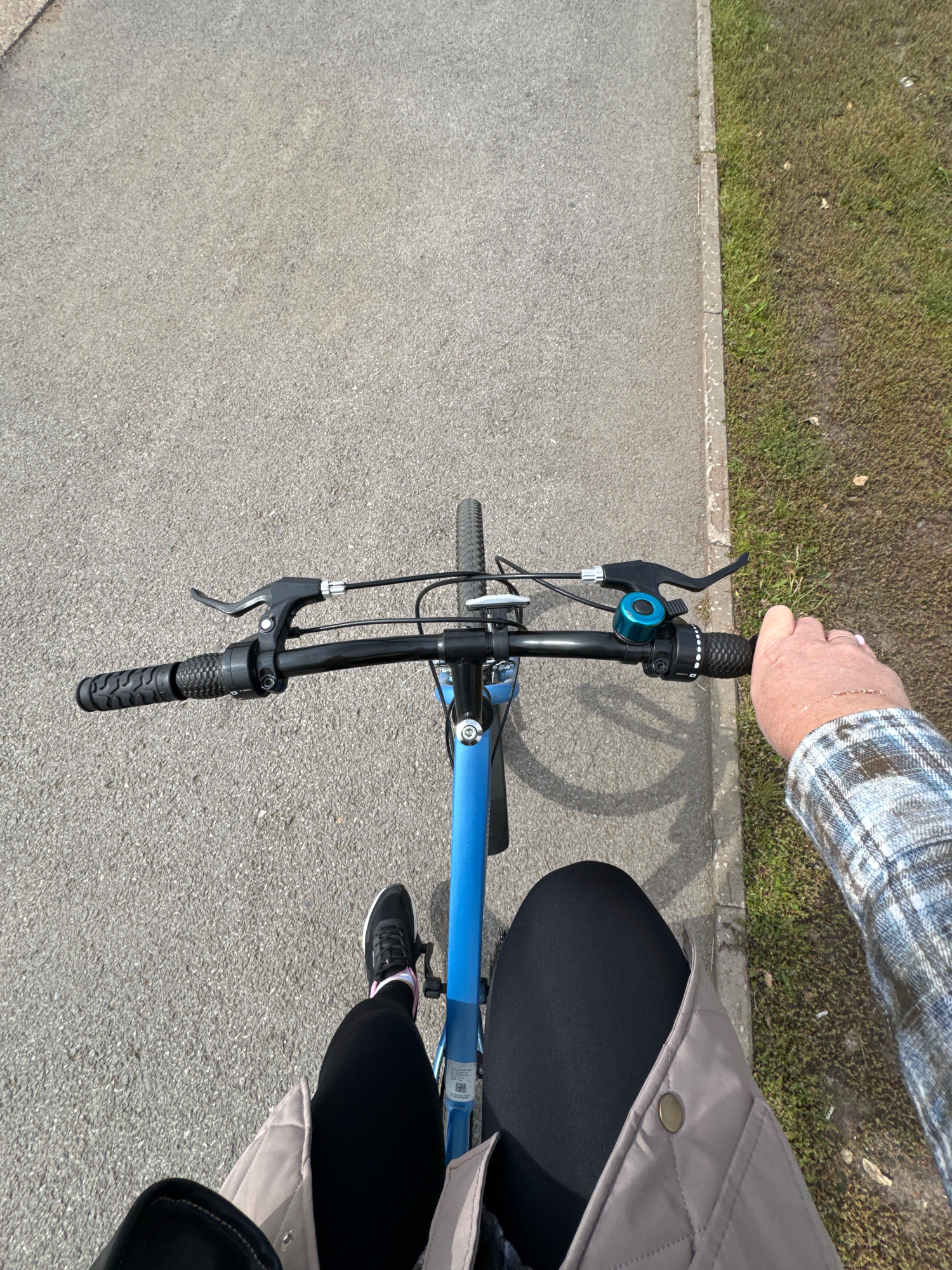 An overhead shot shows a person riding a blue bicycle on a paved path. The path runs alongside a grassy area. The person's legs, clad in black leggings and a grey jacket, are visible. The person is holding the handlebars of the bicycle, which has black grips and a blue bell. A hand wearing a plaid shirt is also visible on the right side of the handlebars. The scene appears to be outdoors on a sunny day. The location of Ufa, Russia is not ascertainable from the visual information provided.