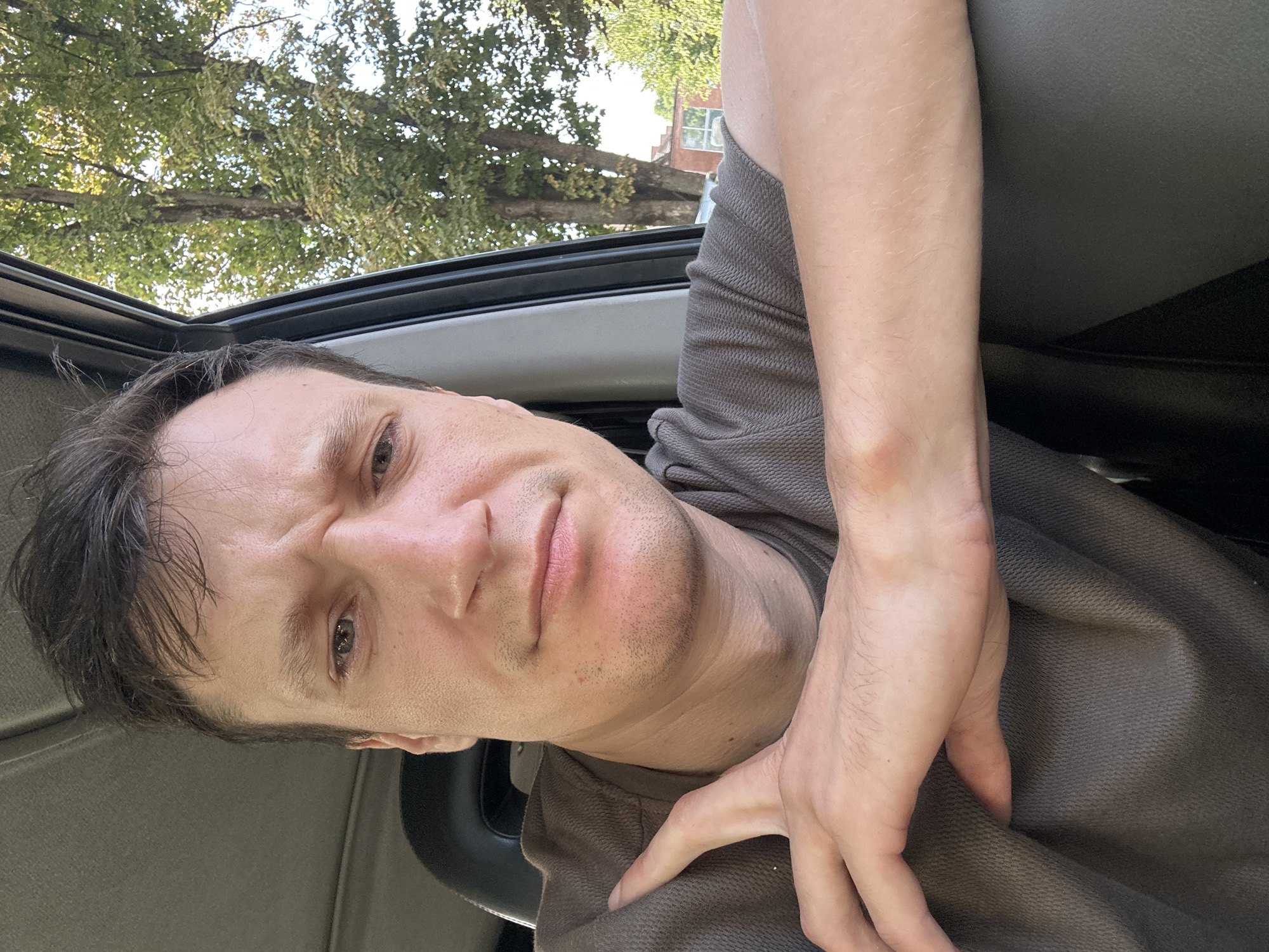 A selfie is taken from inside a vehicle, with the man inside the vehicle. The man is looking towards the camera with a pensive expression, his left arm is resting on the edge of the car door, and his right arm is on his lap. The background shows trees and a building. The image is taken during the daytime, with natural light illuminating the scene.